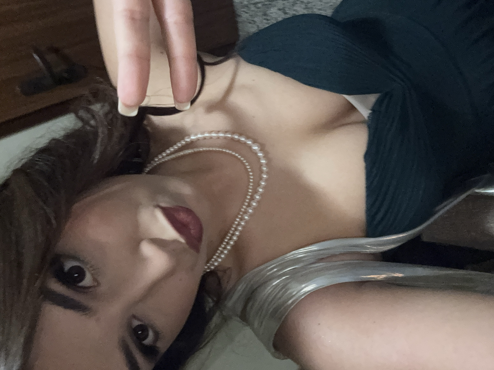 A close-up shot depicts a person wearing a dark green, ribbed dress with a low neckline and a double strand of pearls. The person has dark hair and is wearing dark lipstick and eye makeup. The background is slightly blurred, showing a glimpse of a wooden object and a textured floor. A hand with manicured nails is visible in the upper left corner. A silver-colored material is draped around the person's shoulder or neck. The image is taken at an oblique angle, with the person's face slightly turned towards the camera.
