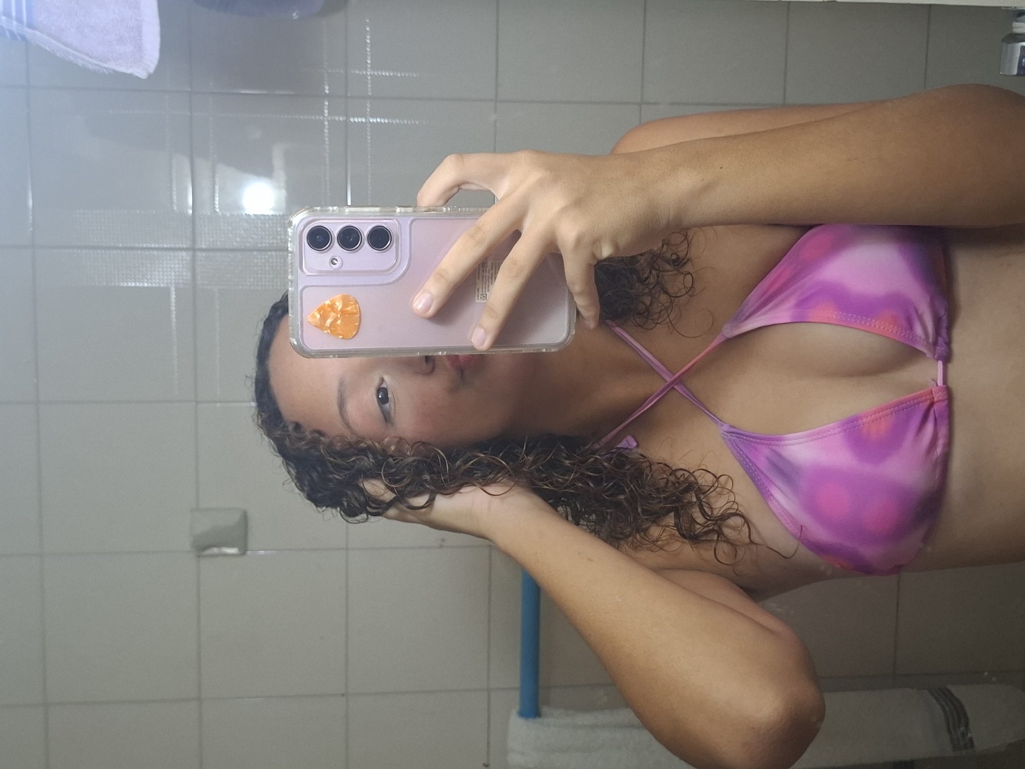 The image shows a person taking a selfie in a bathroom. The person is holding a phone in front of their face, partially obscuring it. They are wearing a pink and purple tie-dye bikini top. The background consists of tiled bathroom walls and a mirror reflecting the scene. The location is Unknown, Unknown.