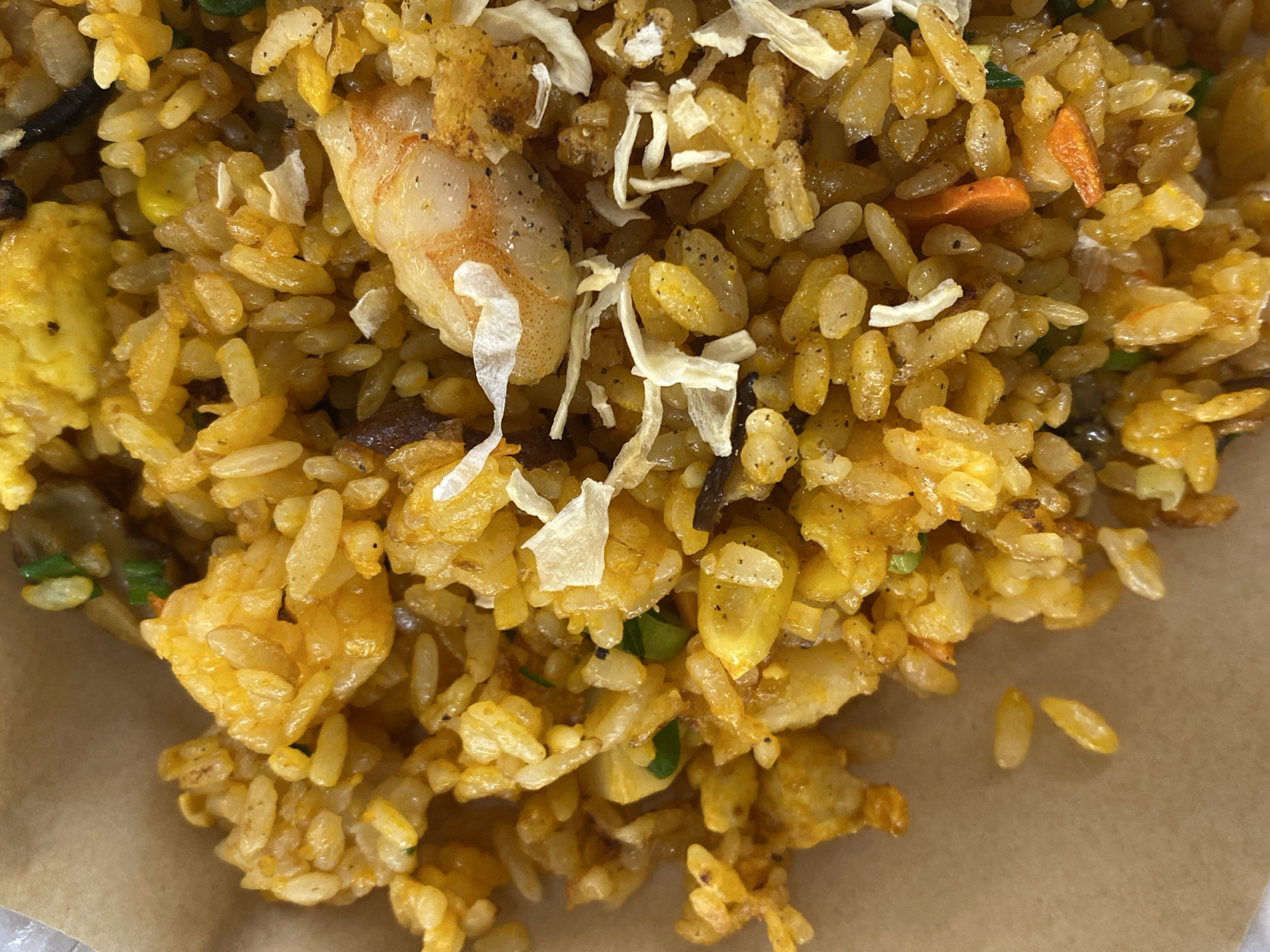 The image shows a close-up, overhead view of a portion of fried rice served in a brown paper container. The rice is a vibrant yellow-orange color, indicating it has been seasoned with turmeric or a similar spice. Visible ingredients include individual grains of rice, pieces of chopped scallions, small diced carrots, and at least one cooked shrimp. There are also some dried, white flakes scattered throughout the dish, possibly dried onion or garlic. The lighting is bright and even, highlighting the textures and colors of the food. The background consists of the brown paper material of the container. There is no visible indication of the location being in Kudrovo, Russia.