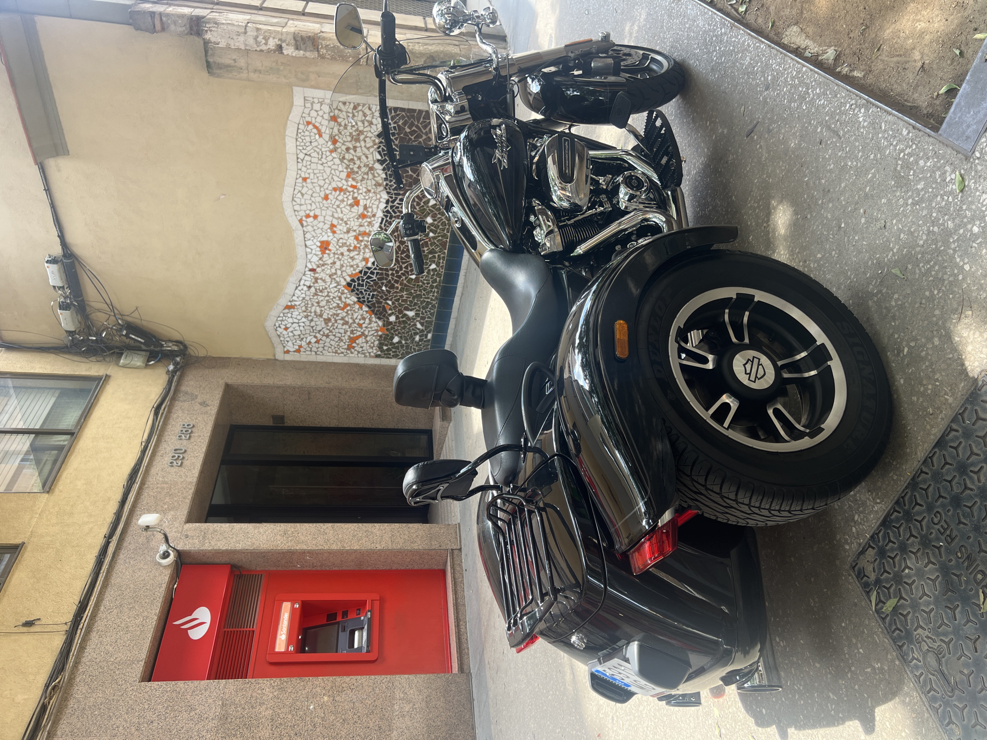 A black Harley Davidson trike motorcycle is parked on a light-colored, textured sidewalk in front of a building in Barcelona, Spain. The building has a yellow-colored facade and a red ATM on the left side, with a "Santander" logo visible on it. Above the ATM, the building number "290 288" is visible. A mosaic art installation runs along the middle section of the building wall behind the motorcycle. The street appears to be a pedestrian walkway. There are no people visible in the image.