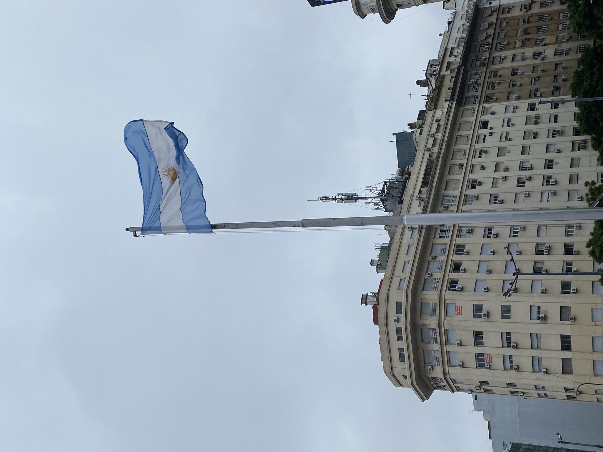 The image shows the flag of Argentina waving in front of a tall building in Buenos Aires. The building has many windows and air conditioning units. The sky is overcast. There are no people visible.