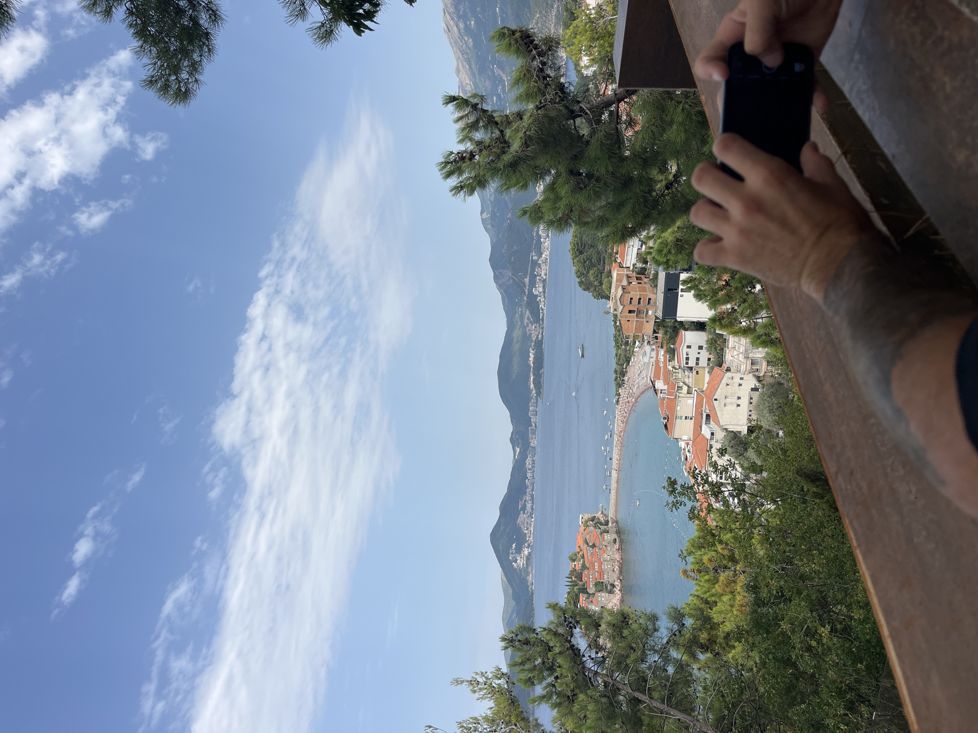 The image shows a scenic view of Sveti Stefan, Montenegro, with the iconic islet in the distance. A person is visible in the foreground, holding a smartphone and taking a picture of the landscape. The islet, connected to the mainland by a narrow causeway, is dotted with red-roofed buildings and surrounded by the clear blue Adriatic Sea. Behind the islet, a crescent-shaped beach is lined with lounge chairs. The background features lush green hills and mountains, with a few boats scattered across the water. The sky is partly cloudy, with a mix of blue sky and white clouds. The foreground is dominated by a wooden railing and the arms of the person taking the photo, who appears to have tattoos on their left arm. The overall scene captures a serene and picturesque coastal setting.
