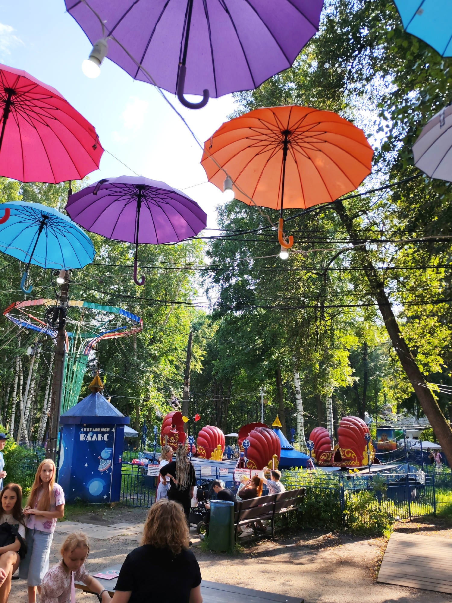 This outdoor scene depicts an amusement park or fairground in Izhevsk, Russia, on a sunny day. Colorful umbrellas are suspended above, adding a festive atmosphere. In the background, several carnival rides are visible, including a carousel-like attraction with red, shell-shaped cars. A blue ticket booth or information stand is present on the left, featuring Cyrillic text and an illustration of a rocket. People are scattered throughout the park, some walking, some sitting on benches, and children are visible playing. The environment is lush with green trees, suggesting a park or wooded area. The overall impression is one of a recreational area filled with activity and vibrant decorations.
