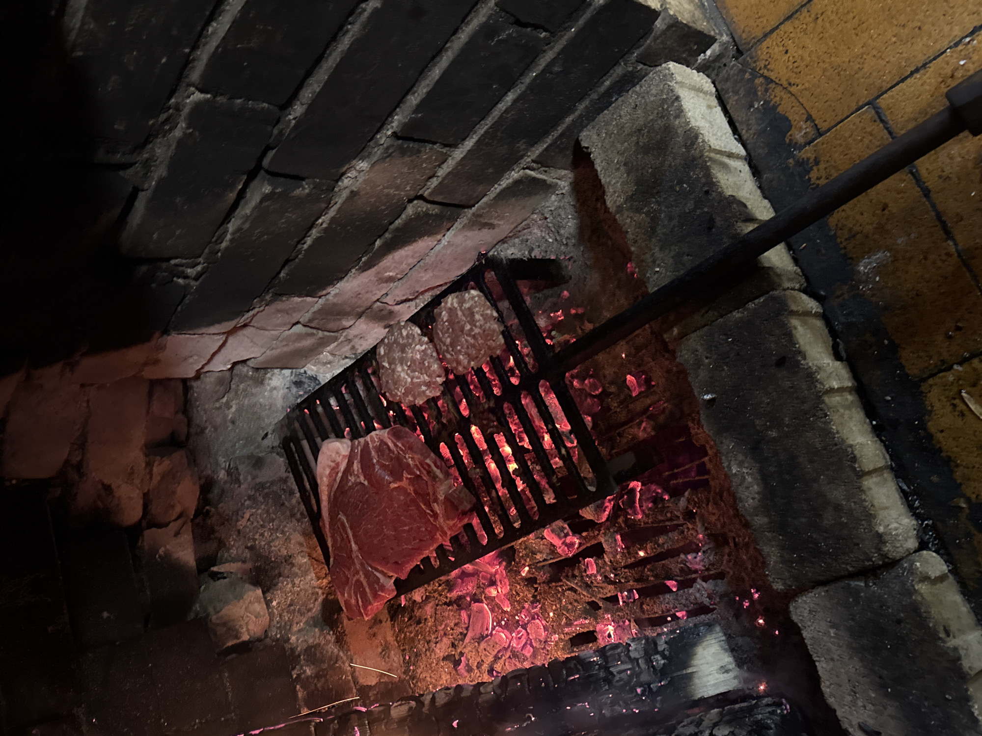 The image shows a grill inside a brick structure, likely a fireplace or oven. The grill contains a large piece of raw meat, possibly a steak, and two smaller circular pieces of meat, possibly burgers or patties. Below the grill are burning coals or embers, creating a red glow and smoke. The brickwork of the structure is visible on the sides and above the grill. A metal rod or handle is visible extending into the scene from the upper right. Location of the scene is unspecified.