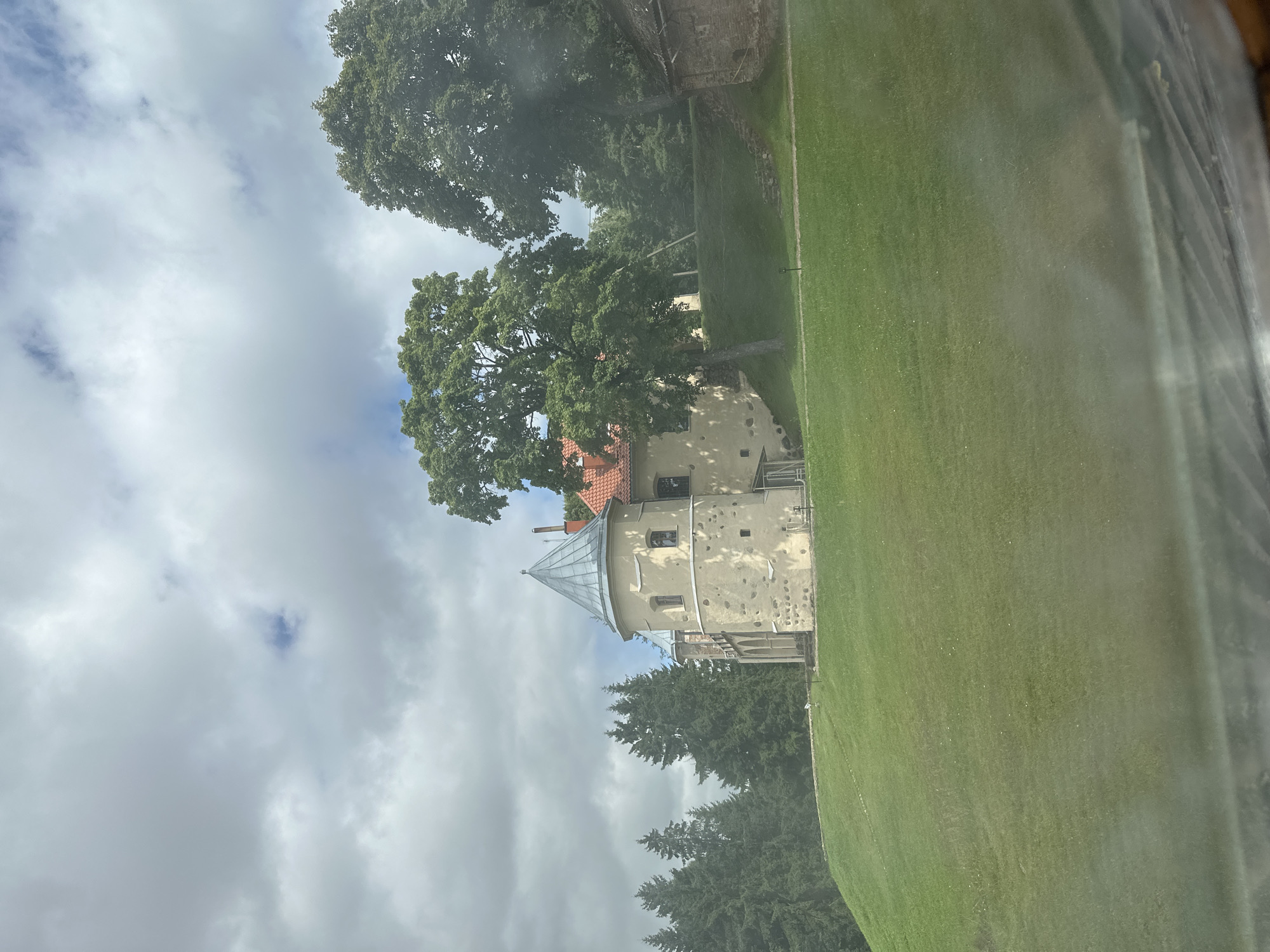 The image shows a stone building with a conical roof, likely a part of Norviliškės Castle in Norviliškės, Lithuania. The building is situated on a grassy hill, with several tall trees surrounding it. The sky is cloudy, with some patches of blue visible. The overall impression is of a historic and picturesque location.