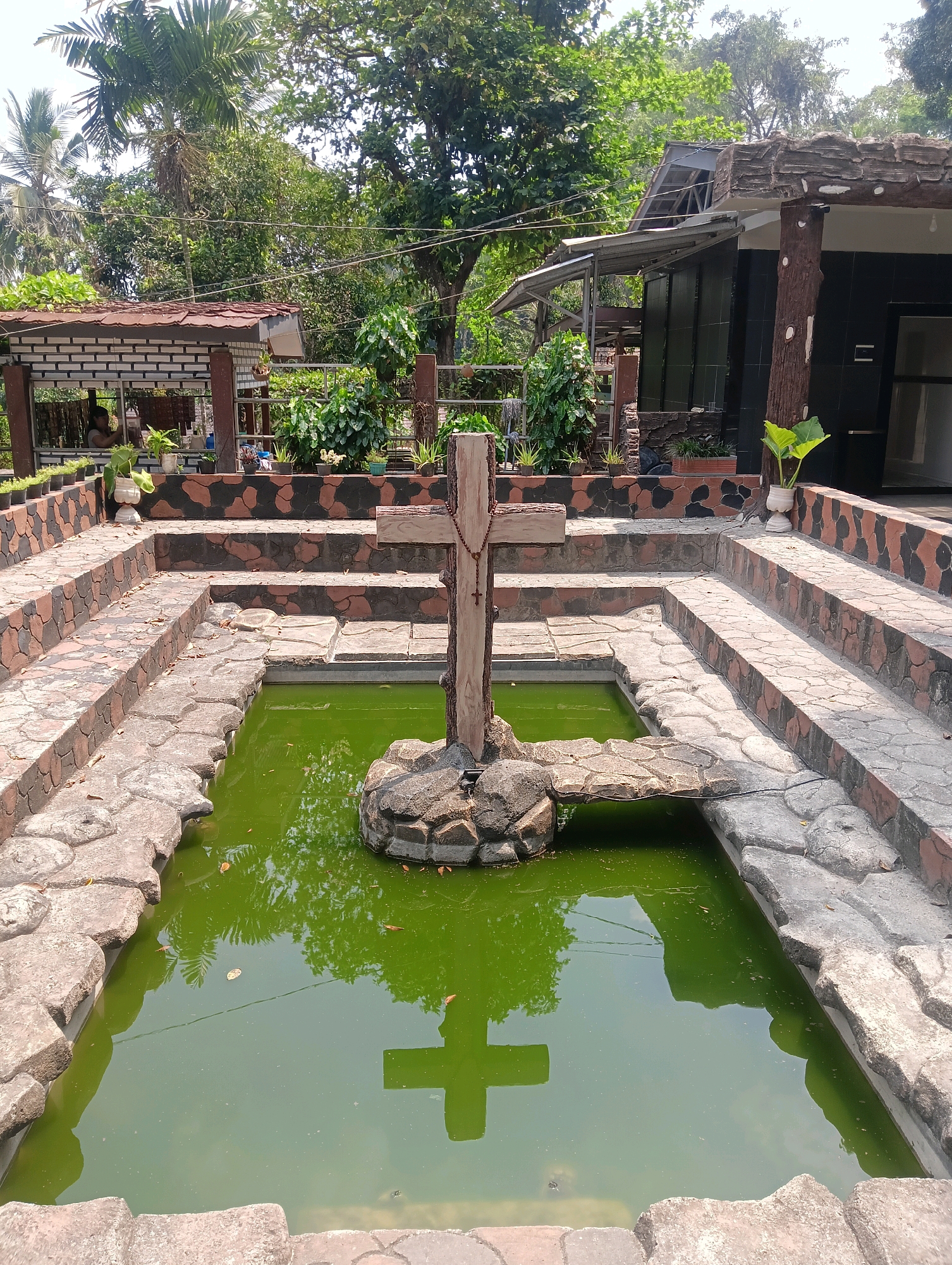 In Narimbang Mulia, Indonesia, a baptismal font is situated outdoors, surrounded by lush greenery and tropical trees. The font itself is a rectangular pool filled with murky green water. A wooden cross, adorned with a rosary, stands prominently in the center of the pool, anchored to a rock formation. The pool is bordered by stone steps and a tiered platform decorated with a camouflage-like pattern of brown and black. In the background, a building with a dark tiled facade and a rustic, wood-like exterior is visible, alongside covered seating areas and potted plants. The overall scene suggests a religious or spiritual site in a tropical setting.
