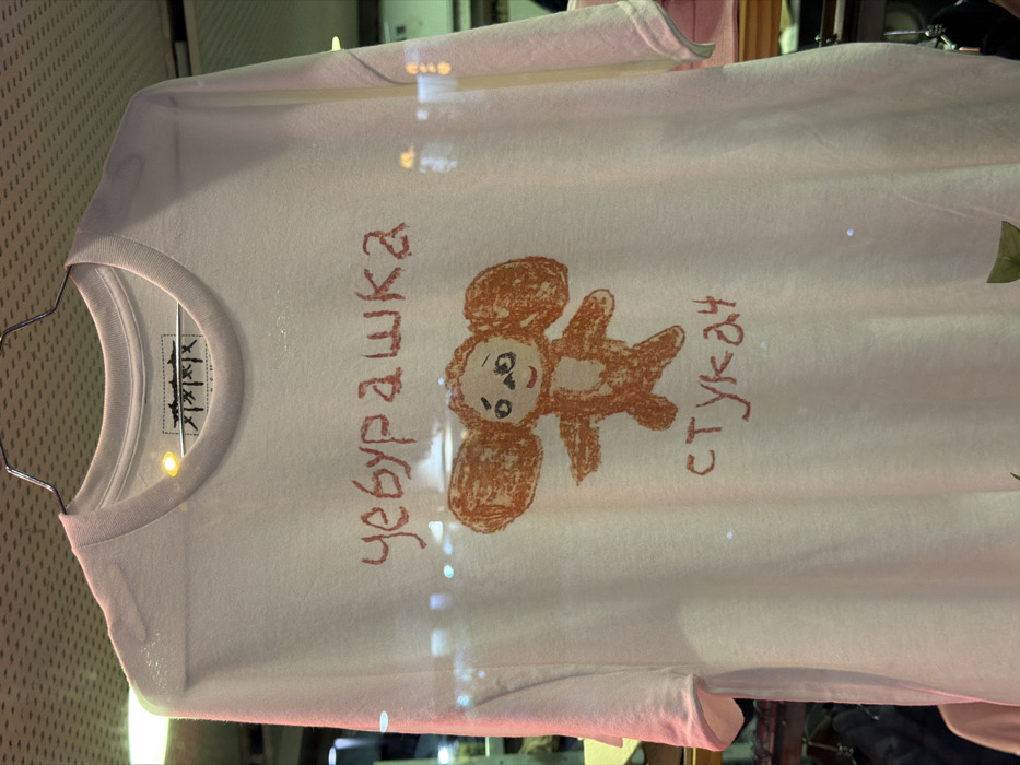 A white t-shirt hangs on a metal hanger, displaying a childlike drawing of Cheburashka in brown crayon. Adjacent to the illustration are the words "Чебурашка" (Cheburashka) and "стукач" (snitch), also written in brown crayon. The shirt's label is visible inside the collar. The lighting and reflections suggest the shirt is displayed behind glass.