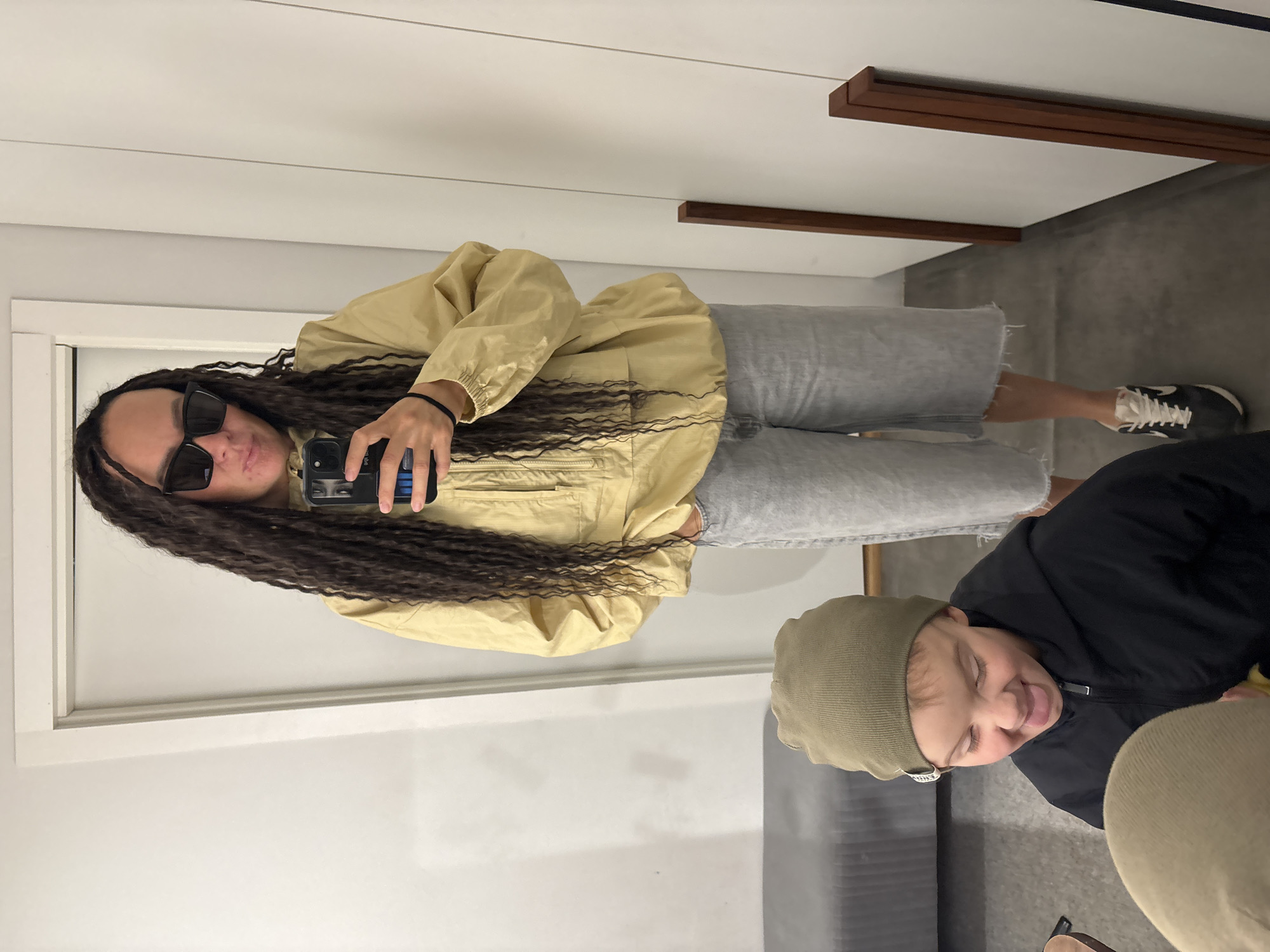 The image, taken in Chaikovsky, Russia, shows an indoor mirror selfie with two individuals. The primary subject, on the left, is a person with long, dark, wavy hair (possibly dreadlocks or braids) that extends past their waist. They are wearing black sunglasses, a light yellow or tan windbreaker-style jacket, and light-wash denim shorts or capris. They hold a dark-colored smartphone with a blue-accented case up to take the selfie, with a black elastic bracelet visible on their wrist. Their expression is obscured by the sunglasses.
In the foreground, on the right, is another person, likely a child, reflected in the mirror. This individual wears a dark jacket and a green/khaki beanie. They are making a playful expression with their eyes squinted or closed and their tongue slightly out.
The setting appears to be a modern interior space. The floor is grey, and a grey upholstered couch or bench is partially visible. The background wall is white, featuring horizontal wooden strips or decorative elements. The primary subject is also wearing black sneakers with white laces and soles, which appear to be Nike shoes based on the visible white swoosh logo on the side. The overall mood is casual and lighthearted.