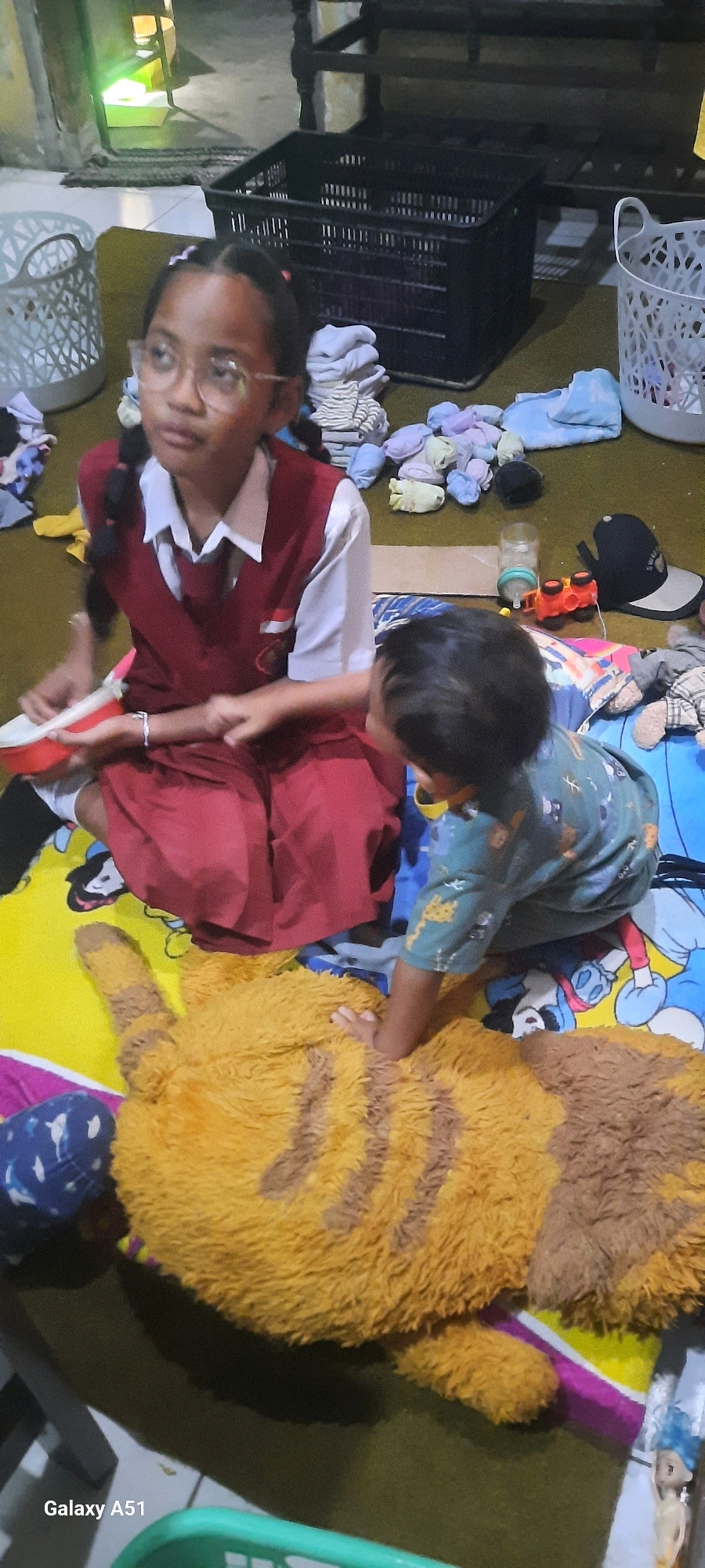 A young girl wearing a red and white school uniform, with glasses and braided hair, sits on a colorful mat. She is holding a red bowl and interacting with a younger child who is reaching towards a large, furry stuffed animal that resembles a tiger. The younger child wears a green shirt with a patterned design. Scattered around them are various items including a black plastic crate, white laundry baskets, stacks of folded clothes, socks, a baby bottle, a toy car, a black baseball cap, and other miscellaneous objects. The scene appears to be indoors, possibly a home, with a tiled floor visible in the background. The lighting suggests it is daytime. The image was captured in Ploso, Indonesia, as indicated by the text "Galaxy A51" in the lower left corner, which is a model of Samsung phone often used in that region.