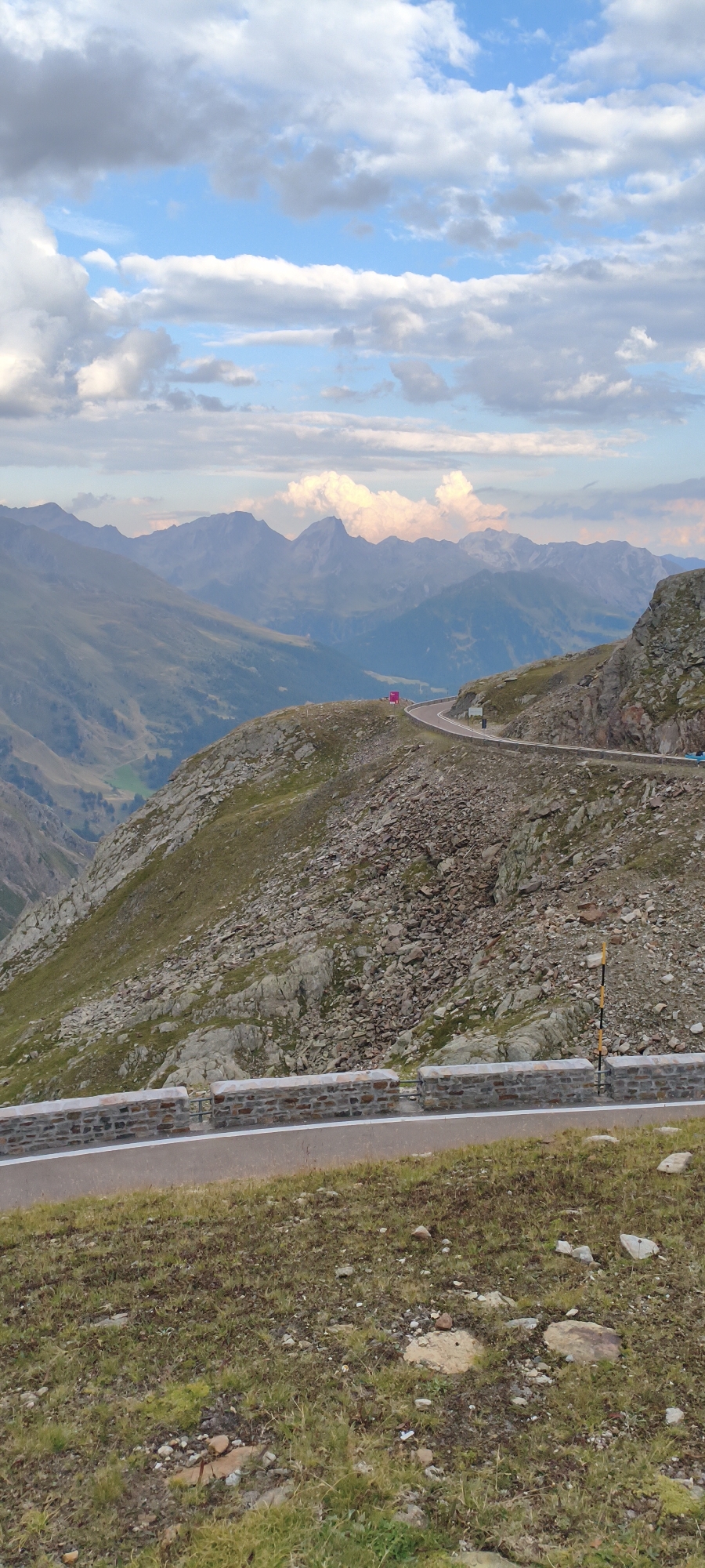 The image shows a winding mountain road in a rocky, sparsely vegetated mountainous terrain. The road has a stone retaining wall along its edge, and white lines mark the lanes. In the distance, layers of mountains recede into the haze under a cloudy sky. There are a few distant vehicles on the road, one of which appears to be a pink structure. The foreground is covered in dry grass and rocks. The scene evokes a sense of elevation and a vast, natural landscape. This appears to be a high mountain pass in the Alps, likely near Sölden, Austria, given the context provided.