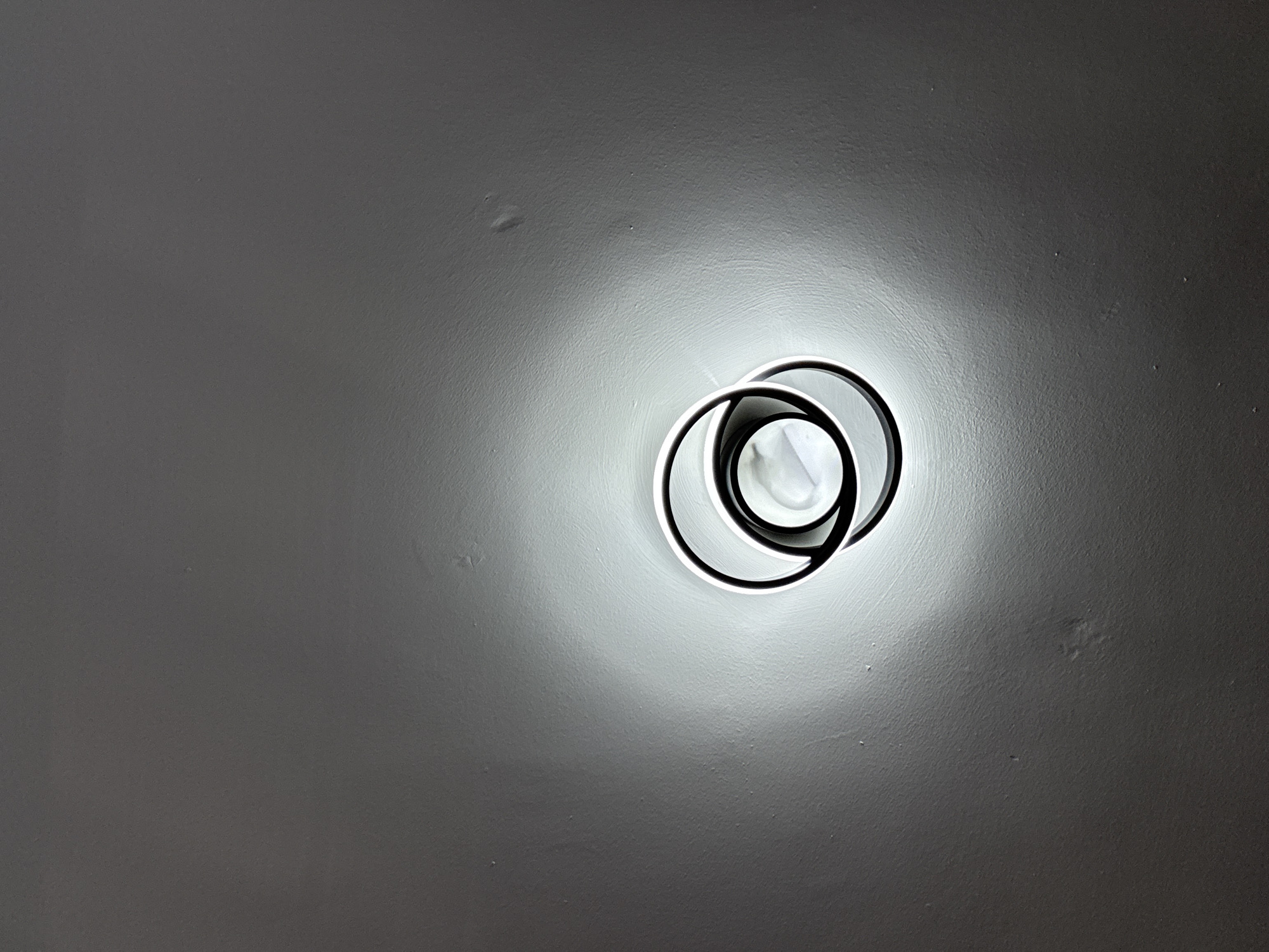 A ceiling-mounted light fixture dominates the center-right of the frame. The fixture consists of overlapping circles, two of which are illuminated, with black accents. The light casts a diffused glow on the surrounding gray ceiling, revealing a textured surface with minor imperfections. The setting appears to be indoors. The location is not discernible from the image.