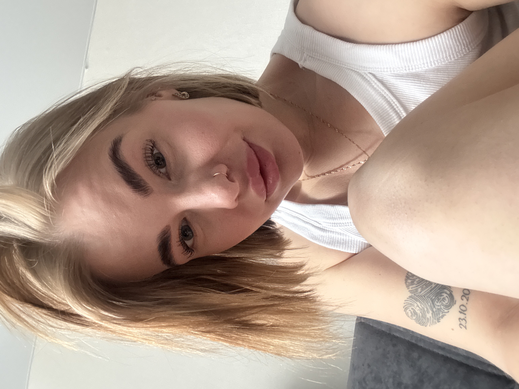 A fair-skinned young woman with blonde, chin-length hair and dark eyebrows poses indoors. She wears a white tank top and a delicate gold necklace. She has a nose ring. A tattoo comprised of fingerprints in the shape of a heart and the numbers "23.10.20" is visible on her arm. The background appears to be a wall and a gray upholstered surface. There is nothing to indicate the specific location in Chaikovsky, Russia.
