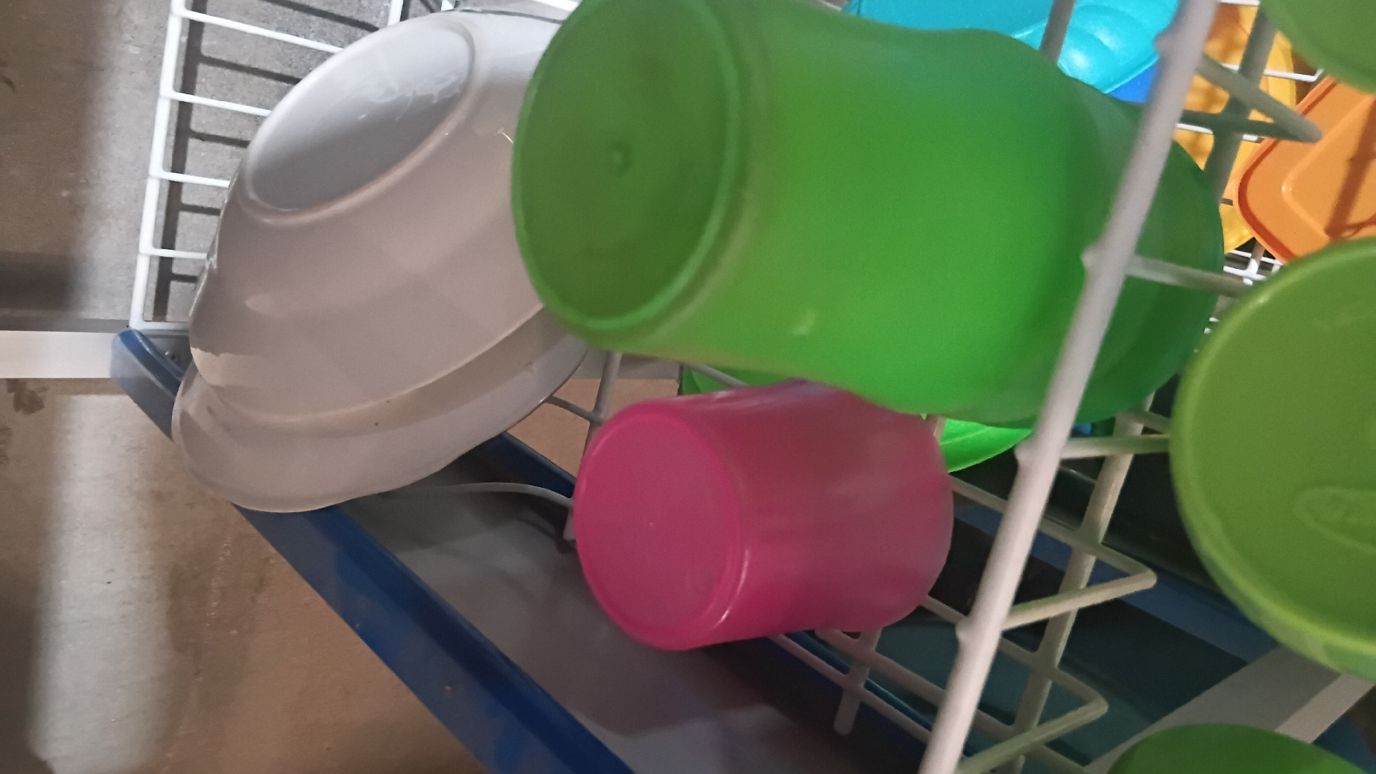 The image shows a dish rack filled with various kitchenware, predominantly plastic cups and bowls. In the foreground, two prominent plastic cups are visible: one is a vibrant pink, and the other is a bright green. Behind them, a large white bowl or plate is stacked, possibly a lid. Other colorful plastic items, including green and orange bowls, are also present in the dish rack. The dish rack itself is white, with a blue rim on the side, and appears to be positioned against a wall with a concrete-like texture. The lighting suggests an indoor setting, possibly a kitchen or utility area. This scene is in the city of Prabumulih, Indonesia.