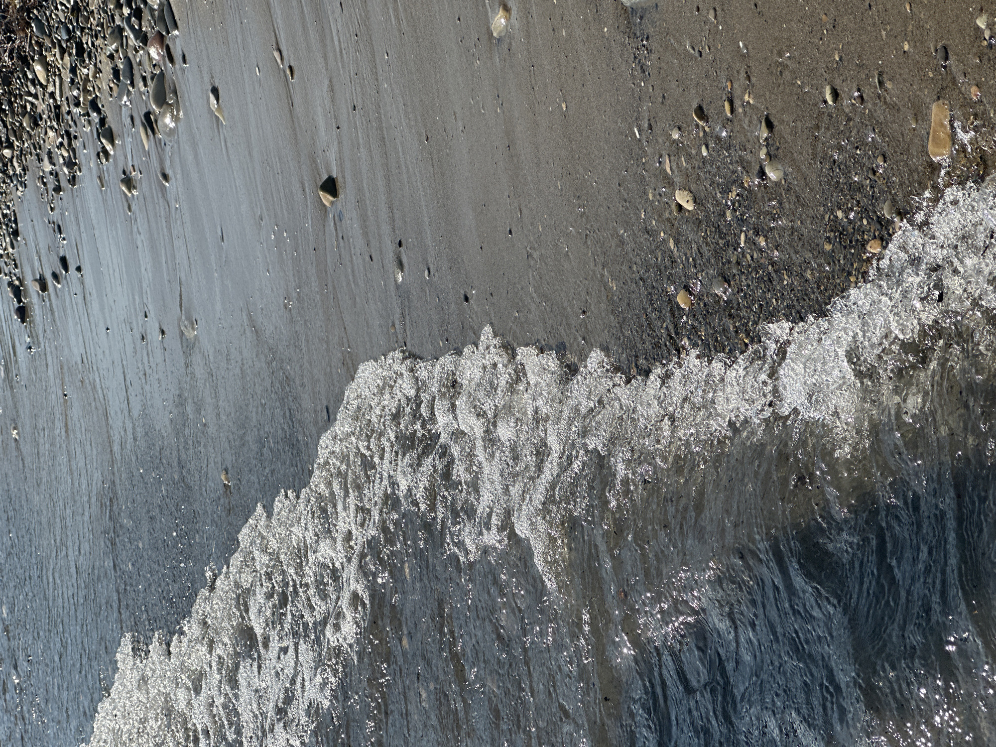 The image presents a high-angle view of a shoreline in Lermontovo, Russia, capturing the immediate interface between water and land under bright illumination.
The scene is diagonally divided by a breaking wave, extending from the upper-left to the lower-right. To the left of this diagonal, the water is a dark, translucent blue-grey, with numerous shimmering reflections of sunlight on its rippled surface, indicating movement and direct sun exposure. The wave itself is a prominent line of white, aerated foam and small splashes, actively cresting as it advances onto the shore.
To the right of the breaking wave, the terrain consists of a sandy and pebbled beach. The sand is a light greyish-tan, appearing darker and smoother where recently wetted by the receding tide, revealing subtle flow patterns. Scattered across the sand are numerous smooth, rounded pebbles and small stones, ranging in size and color, predominantly in shades of grey, brown, and tan. A distinct, somewhat pyramidal stone is visible near the center-right of the sandy area. The density of pebbles increases significantly towards the upper-right corner and along the immediate waterline, where coarser aggregates are present.
The overall environment is natural and devoid of any human presence, subjects, or structures. The lighting suggests a clear, sunny day, with strong highlights on the water and wet surfaces.