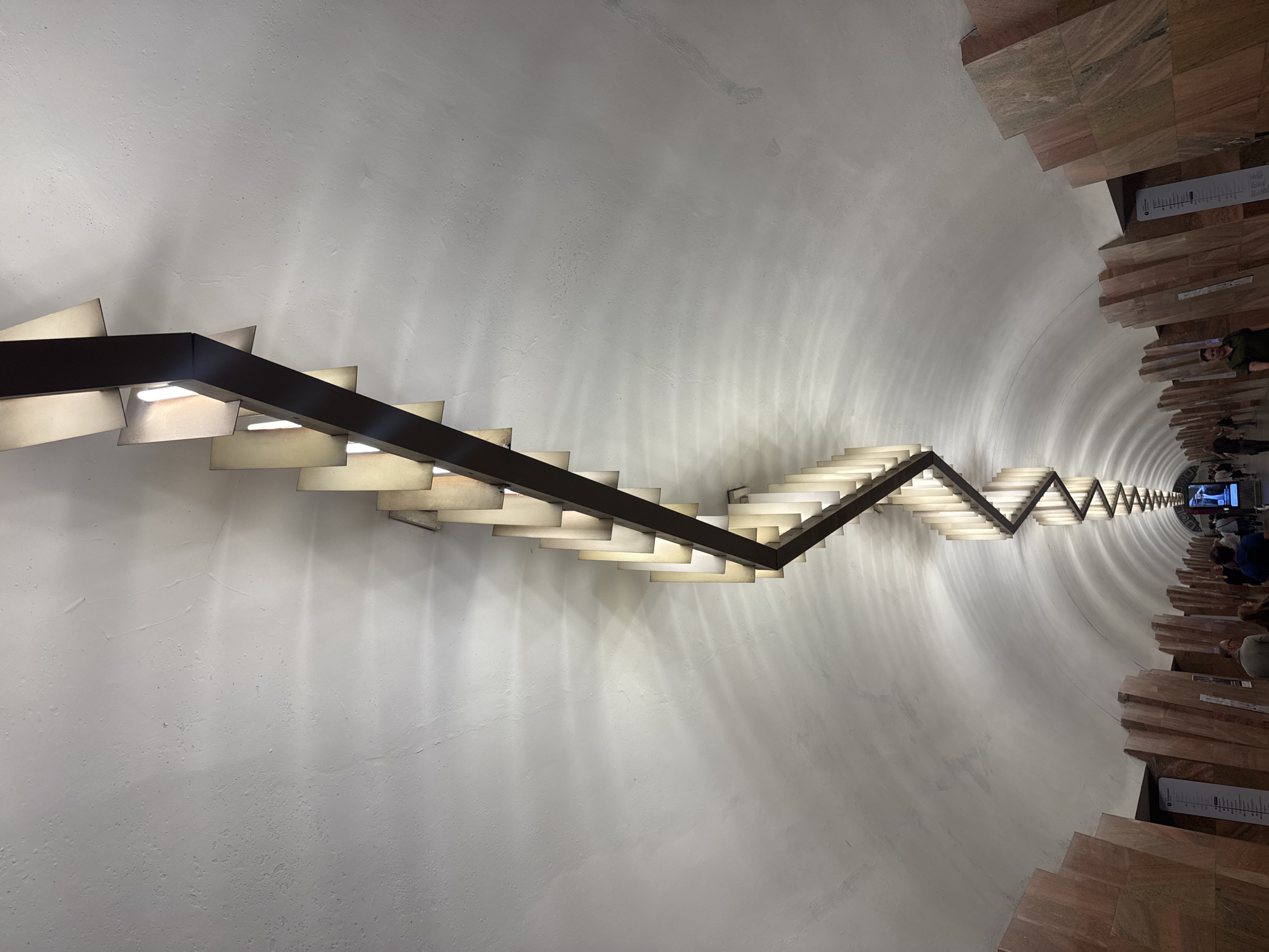 From an elevated perspective, the image captures a distinctive staircase within the Museum of Cosmonautics in Moscow, Russia. The stairwell's design features a cylindrical, slightly elliptical white space containing a series of stairs, each faced with horizontal wooden panels. A zig-zagging dark brown handrail runs through the stairwell, integrated with rectangular light fixtures positioned beneath the rail. The angle of the photograph creates a diminishing perspective, with the stairs becoming increasingly compressed towards the background. A small group of people are visible on the lower stairs, with some gazing towards a display. The overall atmosphere is architectural, emphasizing the spatial design and lighting.