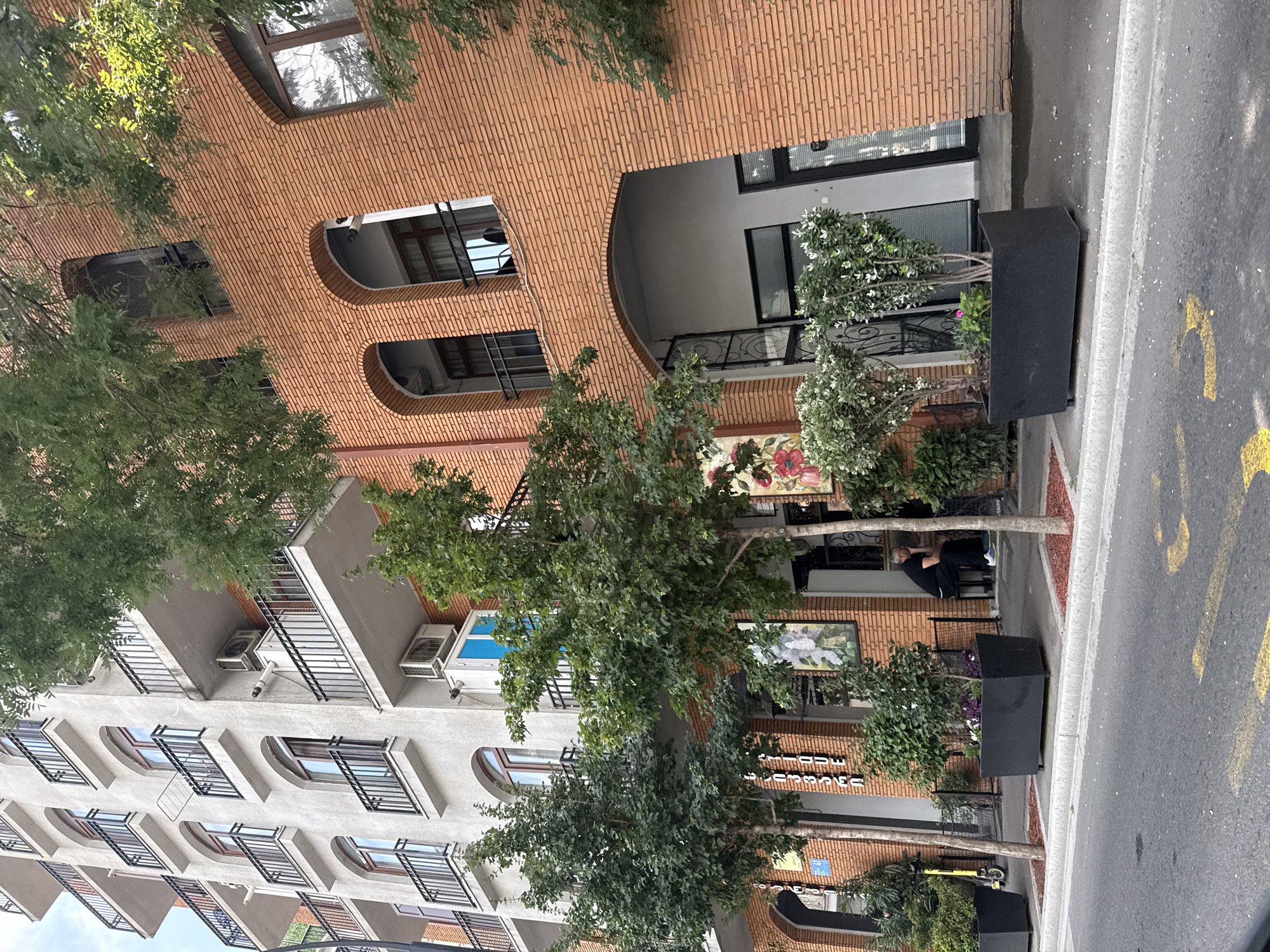 The image shows a street view in Tbilisi, Georgia, featuring a modern building with a light-colored facade on the left and an orange brick building on the right. The buildings have multiple stories with balconies and arched windows. Trees and planters with greenery are situated along the sidewalk in front of the brick building. A person is seated on a bench outside a shop. A yellow electric scooter is parked near the curb. The street is paved, and yellow markings are visible on the asphalt. The sky is partially visible at the top of the image.