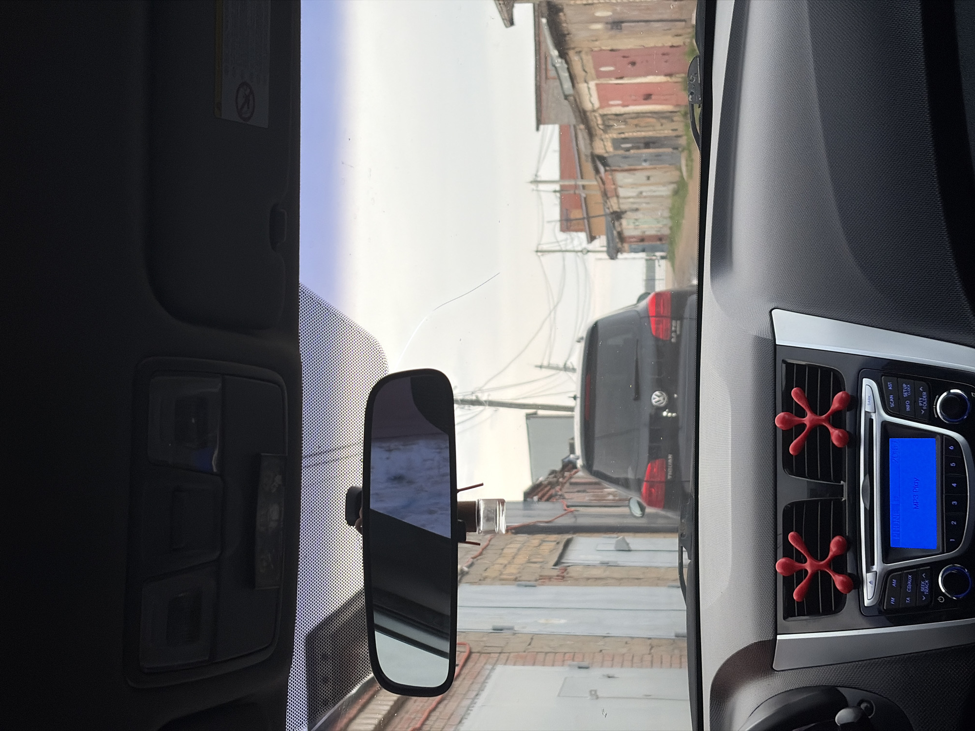 The image shows a view from inside a car, specifically from the driver's seat. The dashboard, windshield, and rearview mirror are visible. In the rearview mirror, the back of a gray Volkswagen minivan can be seen, along with a view of a warehouse and garages. The crack is visible on the upper part of the windshield. The car's dashboard includes a radio with a blue display screen and red, starfish-shaped decorations on the air vents. From the image, it is difficult to identify the exact location in the city of Chaikovsky, Russia.