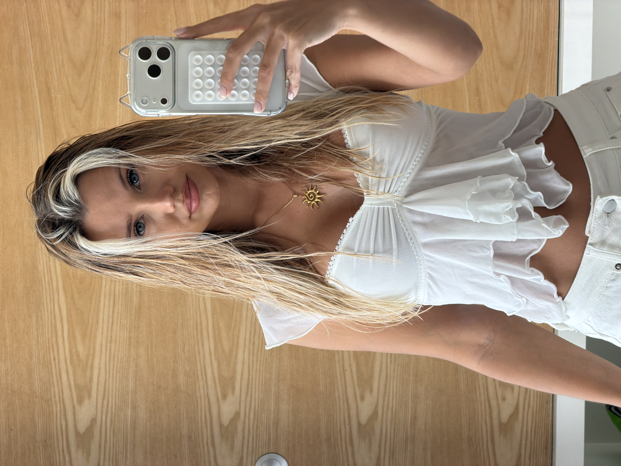 A young woman with blonde hair, featuring prominent platinum blonde streaks, is taking a selfie in a room with light brown wood-paneled walls. She is wearing a white, sheer, ruffled top with delicate lace trim around the neckline, and white bottoms. A gold sun pendant necklace rests on her chest. Her right hand holds a clear phone case with a white textured grip. The background is a plain wood-paneled wall.
