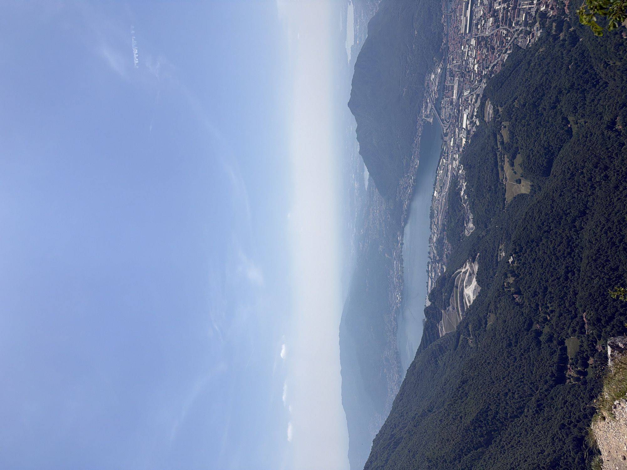 Here's a description of the image:
The image depicts an aerial view of the city of Lecco, Italy, and the surrounding mountainous landscape. The photograph appears to have been taken from a high vantage point, possibly a mountain peak, looking towards the city and Lake Como.
The sky is a pale blue, and there is a haze obscuring the mid-ground.
Lake Como is seen winding between the mountains and the city. The city of Lecco is visible along the shoreline, with its buildings, streets, and industrial areas.
The slopes of the mountains are densely covered with vegetation. A quarry or open mine is visible on one of the slopes.