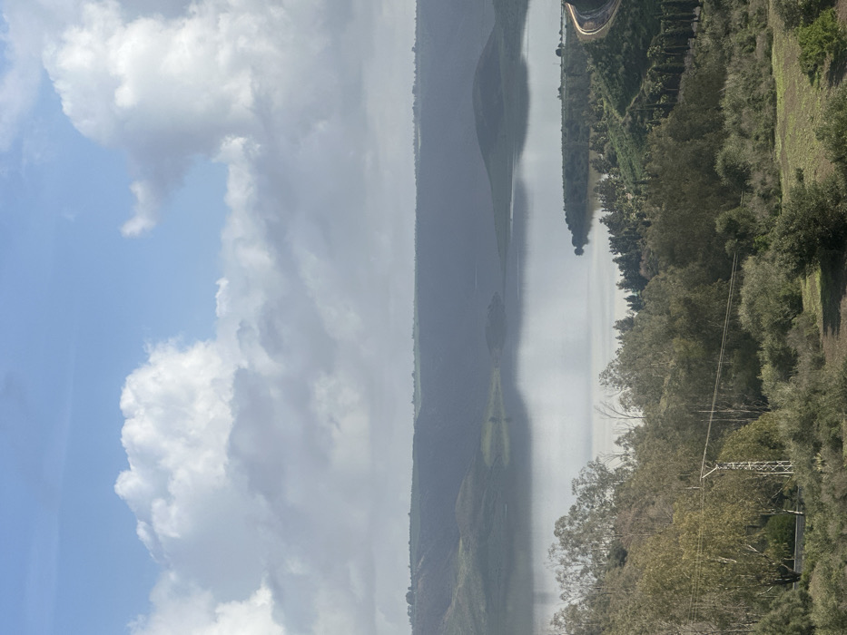 A wide-angle landscape photograph displays a panoramic view of a large body of water, likely a reservoir or lake, situated within a hilly, vegetated environment near Oumazza, Morocco. The sky occupies the upper third of the frame, featuring scattered cumulus clouds against a light blue background; cloud formations vary from bright white to diffuse grey.
The middle ground is dominated by the still, reflective surface of the water, mirroring the sky and distant landforms. Surrounding the water are undulating hills, appearing dark green to bluish-grey due to atmospheric distance, with lighter green cultivated or vegetated patches discernible on their upper slopes.
The foreground and mid-right sections of the image present densely vegetated terrain sloping towards the water's edge. This vegetation includes various green trees and shrubs. A lattice steel electrical utility pole supporting horizontal power lines is visible in the lower-left quadrant. On the far right, a curved segment of a paved road follows the contour of the land adjacent to the water, displaying faint linear light streaks on its surface, potentially from vehicles. The shoreline is irregular, with several small vegetated peninsulas extending into the water. No human figures or animals are present in the observable scene. The environment is under natural daylight illumination.