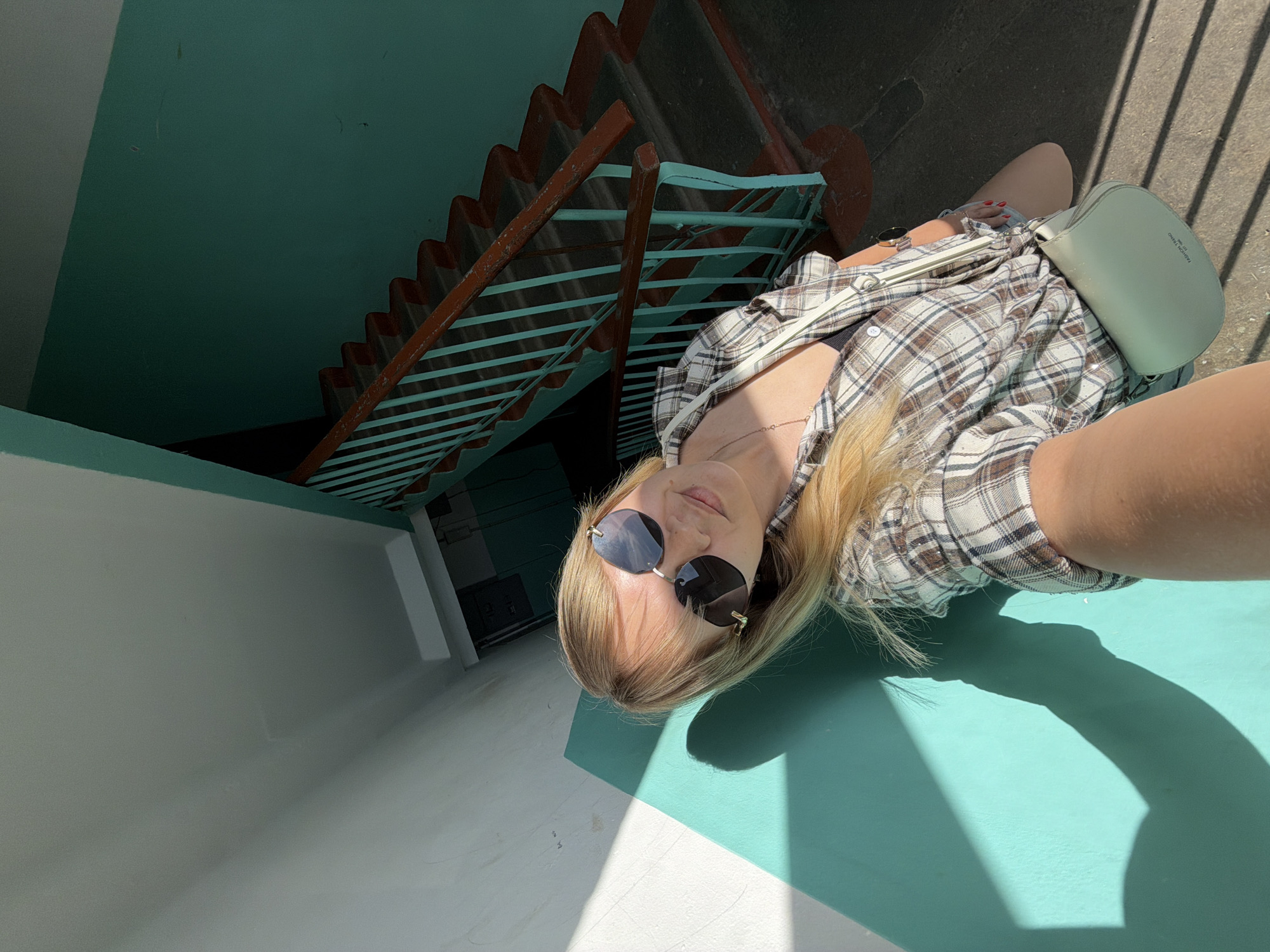 A young woman in sunglasses, a plaid shirt, and a light-colored purse poses in front of a stairwell with teal-colored walls and orange railings. The location is likely Chaikovsky, Russia, based on available information. The stairs lead down into a dark area. Sunlight casts shadows on the teal surface on which she is lying.