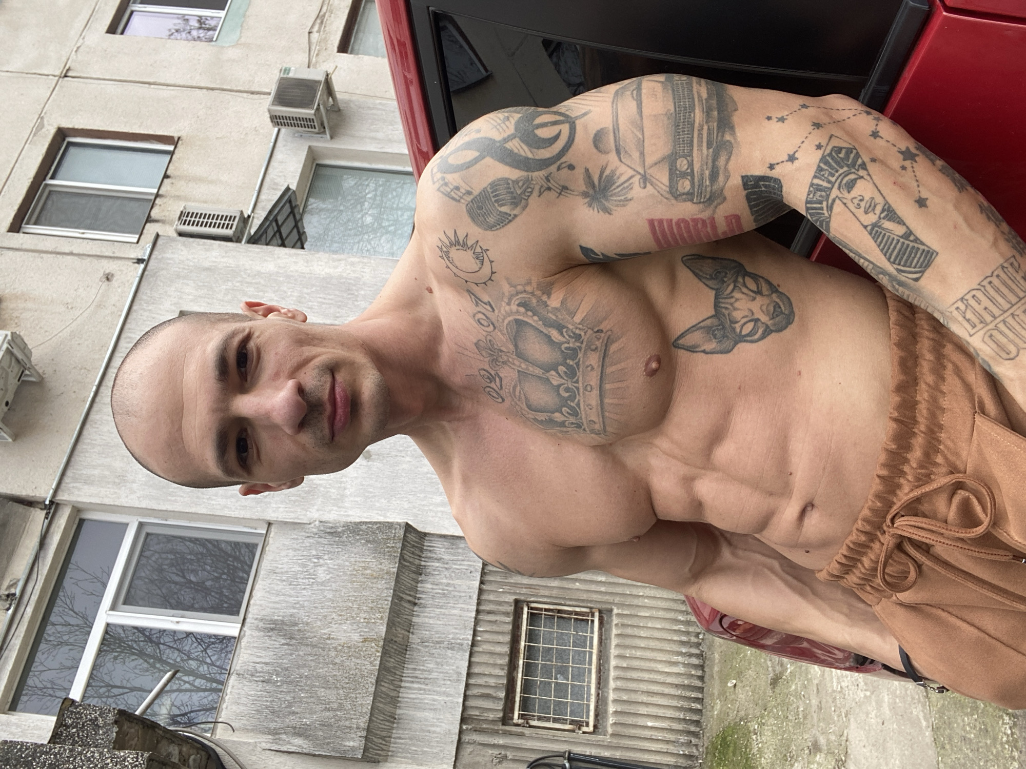 A shirtless man with tattoos stands outside in front of a building. The man is muscular with a shaved head. He has multiple tattoos on his arms, chest, and torso, including images of a microphone, a cat, a crown, and the word "WORLD". He is wearing brown pants with a drawstring. He is standing next to a red car. Behind him is a weathered, multi-story building with windows and air conditioning units. Based on the architectural style of the building and its location near the person, it is likely in the city of Pleven, Bulgaria. The image appears to be a selfie taken from a low angle.