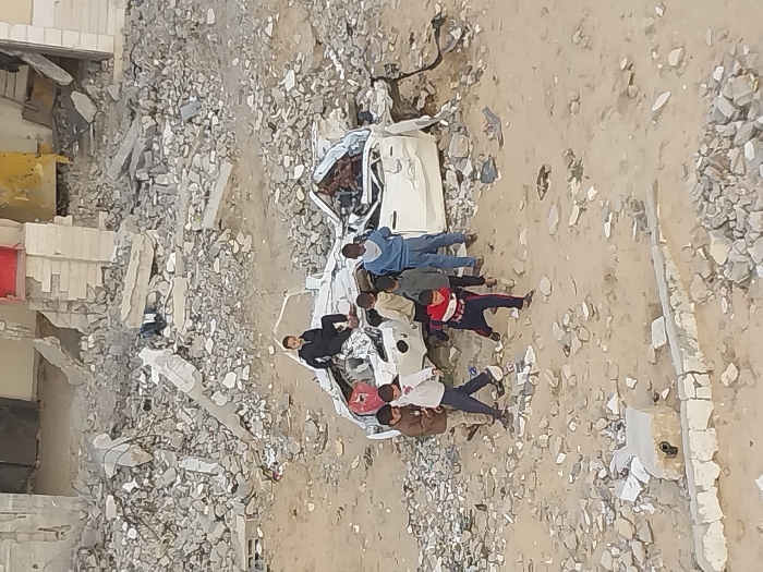 The image depicts a scene of extensive destruction in Khan Yunis, Palestinian Territory. The environment is dominated by vast amounts of building rubble, broken concrete, and debris scattered across sandy ground. In the background, partially destroyed structures are visible, with exposed internal components and collapsed sections; a red door and a yellow wall fragment are distinguishable on one building.
Centrally positioned is a severely damaged white vehicle, likely a car, with its roof caved in, body panels crumpled, and significant structural compromise to its front end. This vehicle appears to be an immobile wreck.
Seven children are present in the scene. One child, dressed in a dark tracksuit, is seated on the mangled roof of the white car. The other six children are standing or moving around the car wreckage and the surrounding debris. One child in a red and dark blue tracksuit is captured mid-stride in the foreground, seemingly running or walking quickly. The remaining children are distributed near the car, observing or interacting with their surroundings. Their actions suggest an informal exploration of the debris-strewn landscape. In the immediate foreground, a damaged low concrete wall or curb is visible amidst the rubble.