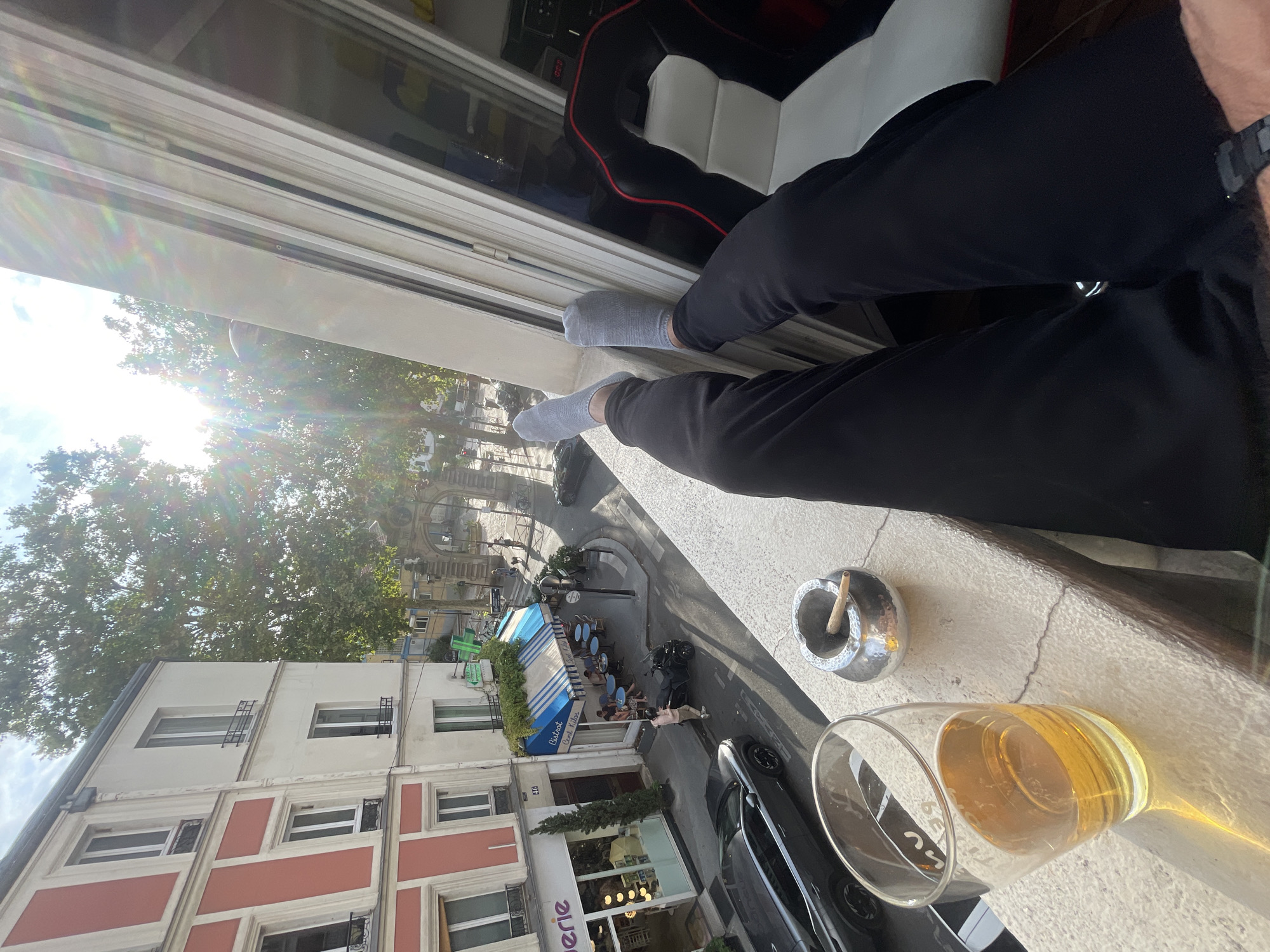 A first-person perspective shot shows a person's legs and feet clad in grey socks and black pants, resting on a balcony railing overlooking a Parisian street scene. To the left of the legs is a glass of amber liquid, likely beer, and a metal ashtray containing a lit cigarette. Below the balcony, a street with parked cars, a motorcycle, and pedestrians unfolds. A restaurant with blue and white striped awnings and outdoor seating is visible, as is a building with red and cream colored facade. The sun shines brightly, casting long shadows and creating lens flare. The scene is likely a residential area in Paris, given the architecture and the presence of a cafe. The overall mood is relaxed and leisurely.