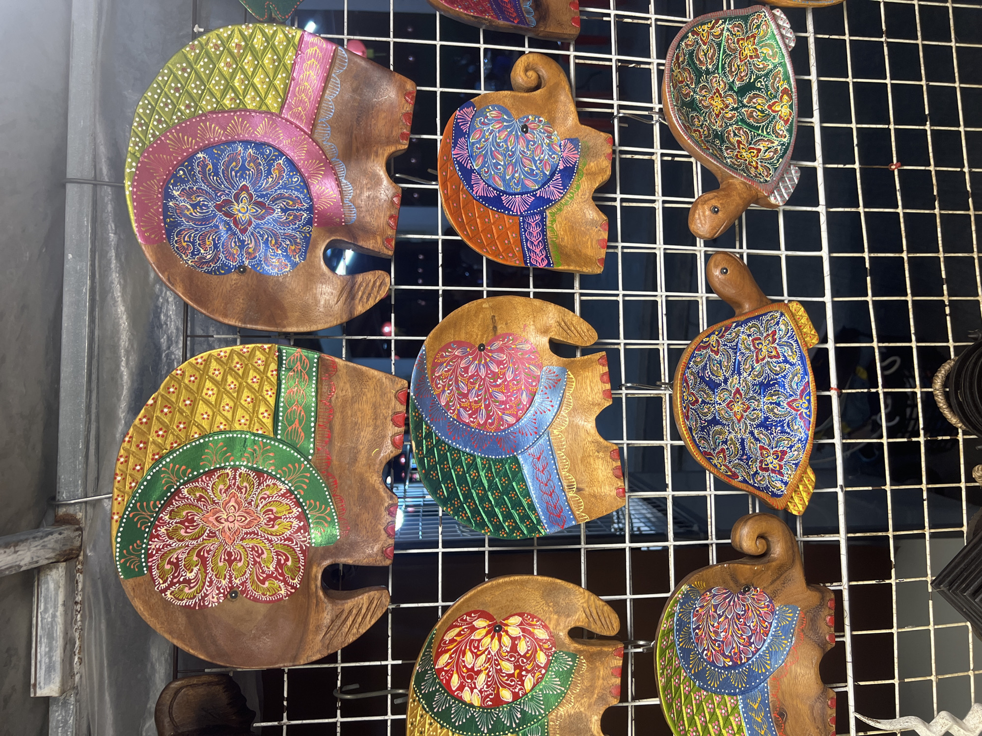 The image displays a collection of vibrantly painted wooden animal figures, primarily elephants and turtles, hanging on a white metal grid. The figures are intricately decorated with colorful, paisley-like patterns in shades of red, pink, blue, green, and yellow. The background consists of a dark, indistinct wall or display, with some metal shelving and lighting visible at the top of the frame. The overall scene suggests a market stall or shop selling handicrafts, likely in a tourist area. The context of ตำบลเกาะพะงัน, Thailand, implies this could be a souvenir shop on the island of Koh Phangan, known for its markets and artisanal crafts.