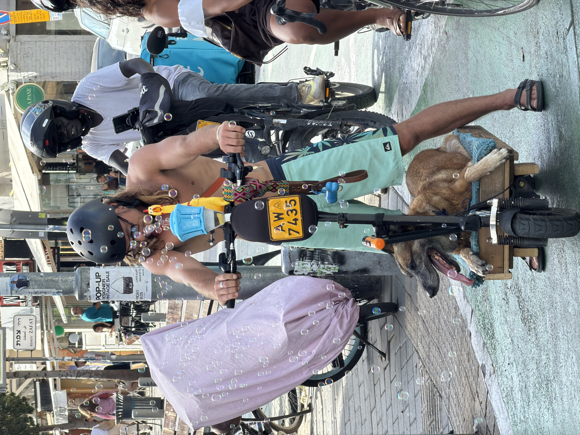 The image depicts a street scene in Tel-Aviv, Israel. A man wearing a helmet and beach shorts is riding a bicycle with a dog in a wooden crate attached to the front. The bicycle has a license plate with the number AW 7435. Bubbles are surrounding the man and bike. In the background, there are pedestrians, parked cars, and a scooter. There are signs for a "Pop-Up Designer Vintage Sale" and a restaurant called "A Pane". A sign reading "REHOV ZAFAT" is also visible, indicating the street name. Another man wearing a helmet is behind the man riding the bike.