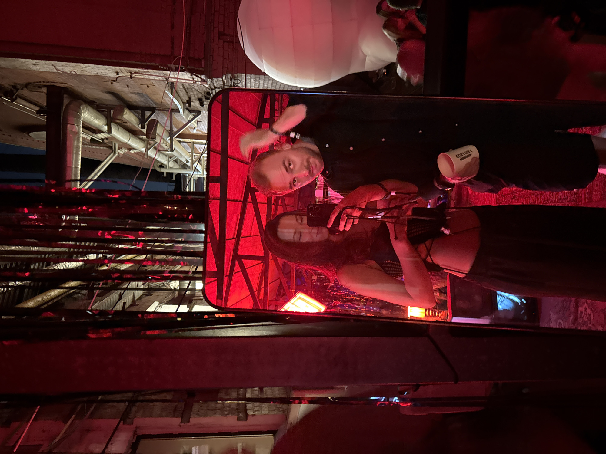 The image depicts two individuals reflected in a large, dark-framed mirror with a rounded top, set within an industrial-style environment illuminated predominantly by red light.
In the mirror's reflection, on the left, a woman with long dark hair is holding a dark-colored smartphone, likely taking a selfie or video. She wears a black mesh or lace crop top and dark, loose-fitting trousers or a skirt, exposing her midriff. Her face is partially obscured by the phone. On the right, a man with short brown hair and light stubble is looking towards the phone, his left hand touching his head. He wears a dark, possibly black, button-up shirt and dark shorts, holding a white ceramic mug with visible dark text (partially decipherable as "DOVE") in his right hand.
The immediate background outside the mirror, particularly to the left, shows a dark structural element or pillar. Above and to the right of the mirror, vertical red strips hang down, creating a reflective curtain-like effect. The broader environment reveals an exposed brick wall, multiple insulated and uninsulated metal pipes running along it, and structural beams, indicating an old factory or industrial building. In the upper right corner, a glimpse of a dark sky suggests an outdoor or open-air space. To the far right, behind the man's reflection, a large white inflatable object, abstract in shape, is visible, along with blurred figures suggesting a crowd. A small, orange-glowing lamp is visible within the mirror's reflection, resting on a dark console or cabinet. The entire scene is bathed in a deep red ambient light, suggesting a club, event, or art installation.
Based on the distinctive industrial architecture, exposed piping, brickwork, and the vibrant, red-lit event atmosphere, the location is characteristic of renovated industrial spaces used for creative events, art installations, or nightlife venues in Moscow, Russia. Such aesthetics are notably found in districts like Artplay, Flacon Design Factory, or specific club venues that repurposed old factory buildings.