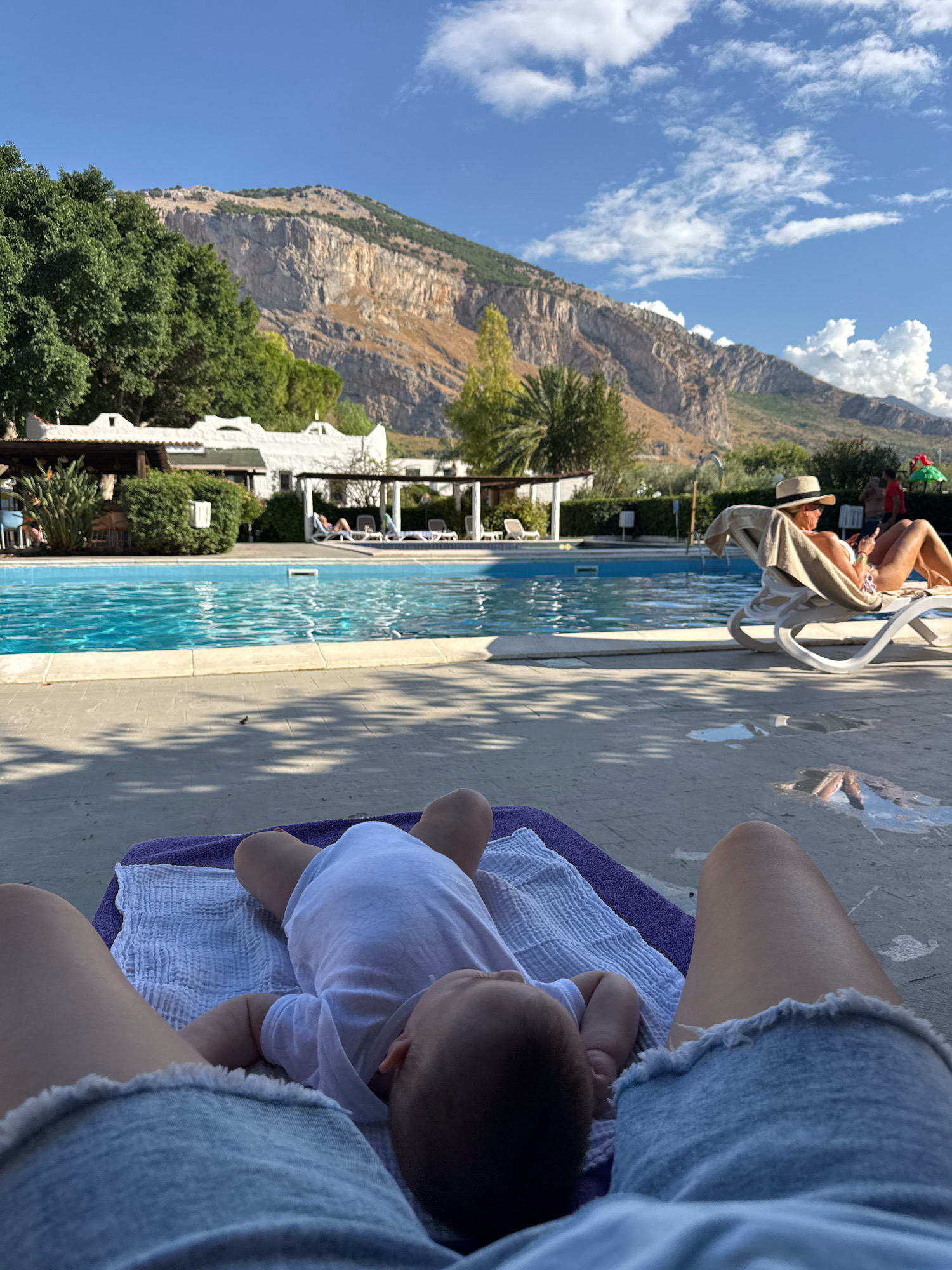 This image depicts a sunny outdoor scene at what appears to be a resort or hotel with a swimming pool. In the foreground, a baby lies on its back on a purple mat and white towels, wearing a white onesie. Next to the baby are the legs of an adult wearing denim shorts.
In the mid-ground, a bright blue swimming pool stretches across the frame. The pool area is paved with concrete, and several white lounge chairs are visible, some occupied. A woman wearing a wide-brimmed hat is relaxing on a lounge chair, holding a phone. Other individuals are visible in the background near the pool, and a building with white awnings is situated behind the pool, surrounded by greenery.
The background features a dramatic mountainous landscape under a clear blue sky with scattered white clouds. Lush green trees and some palm trees are scattered throughout the scene. The overall impression is one of a relaxed holiday atmosphere.
The image does not provide specific visual cues to pinpoint the exact location within Isola delle Femmine, Italy. However, the presence of a pool, resort-like buildings, and a Mediterranean mountainous backdrop is consistent with the geography of coastal towns in that region.