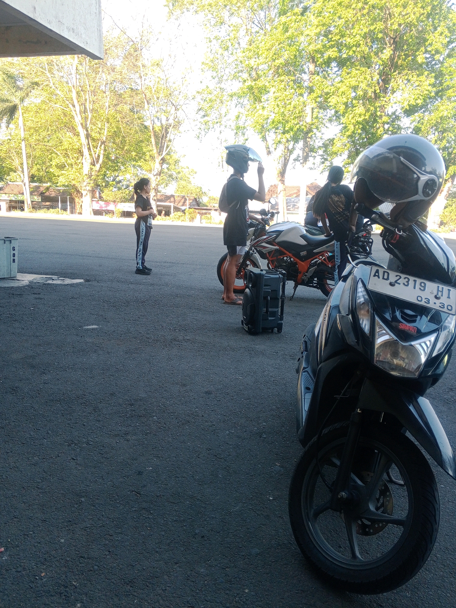 In Wuryorejo, Indonesia, two individuals, one a young woman with dark hair pulled back and wearing a black t-shirt and striped athletic pants, stands holding a water bottle and looking to the right. Beside her, another person, also wearing a helmet and a black t-shirt, is adjusting their helmet and facing away from the camera. A black, wheeled carrying case is positioned on the asphalt ground. In the foreground, a black scooter is parked, its license plate reading "AD 2319 HI 03.30". Behind the scooter and individuals, another motorcycle, predominantly white with orange accents, is visible. The scene is set outdoors with paved ground and a backdrop of trees. A concrete structure and a garbage bin marked "GOR" are also present on the left side of the image. The lighting suggests it is daytime, likely in the morning or late afternoon.