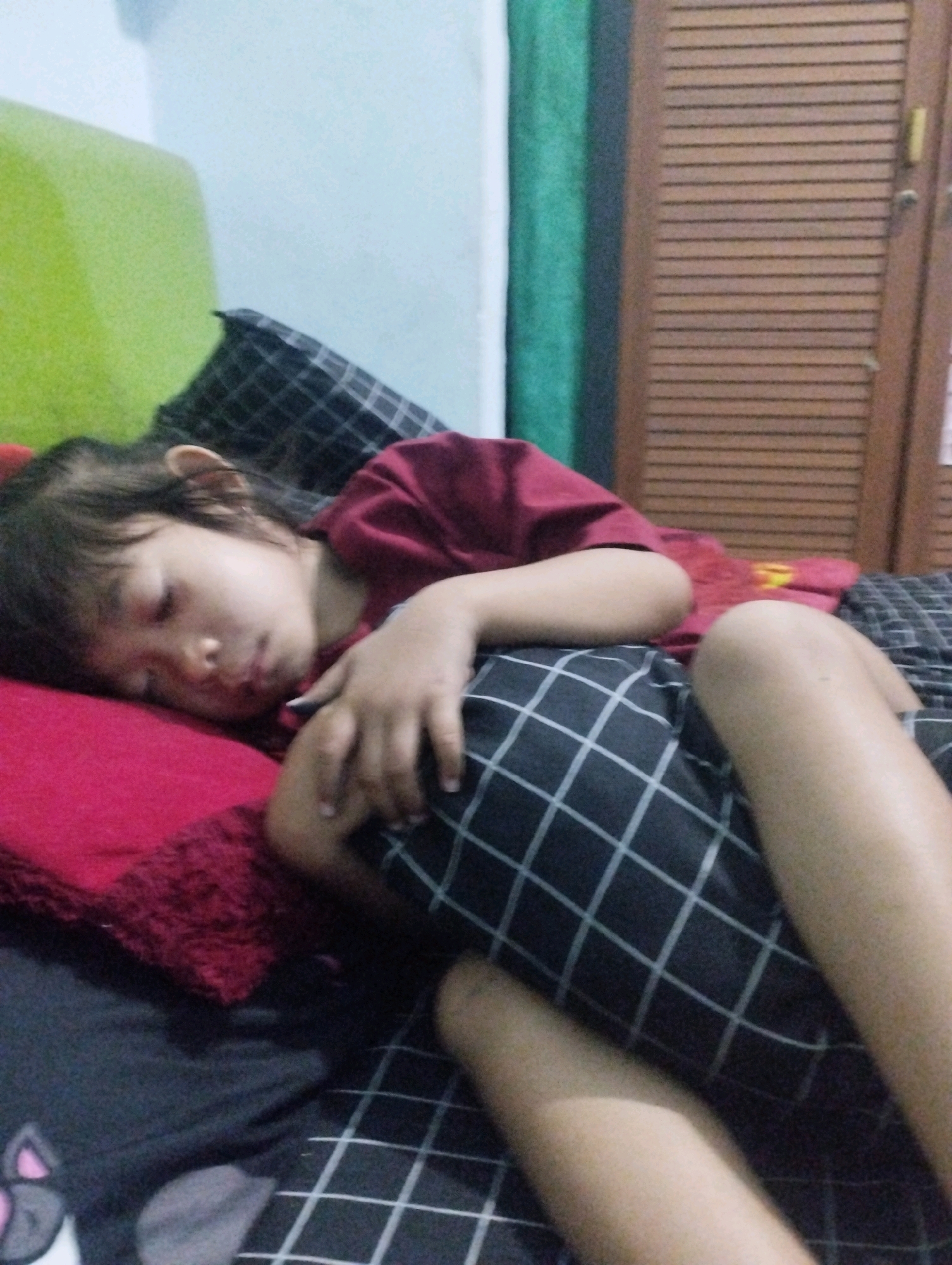 A child sleeps soundly on a bed. The child is lying on their side, with their face turned away from the camera, resting on a red pillow. They are wearing a maroon t-shirt and black and white checkered shorts. The bed appears to be in a bedroom, with a green headboard visible in the background, and a wooden wardrobe with slatted doors on the right side. A dark grey blanket is partially visible at the bottom of the frame. The image is taken indoors, likely in a residential setting in Kemantren, Indonesia, as suggested by the overall domestic environment.