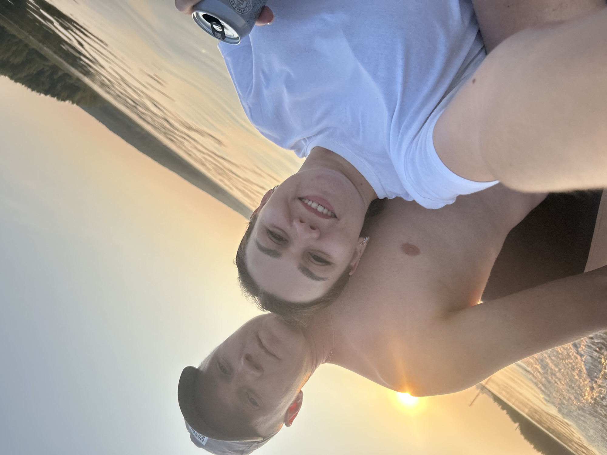The image shows two people on a body of water, likely a lake or river, with a wooded shoreline in the background. The sun is low in the sky, casting a warm, golden glow, indicating it is either sunrise or sunset. The water is calm, with ripples reflecting the light. The person in the foreground is wearing a white t-shirt and is holding a can. The second person is wearing a baseball cap. They appear to be taking a selfie, looking towards the camera with smiles. The overall mood is relaxed and happy, suggesting a leisure activity like boating or swimming during a summer evening or morning. No visible text is present on the people or objects, other than what might be on the can, which is partially obscured. The location is given as Chaykovsky, Russia.