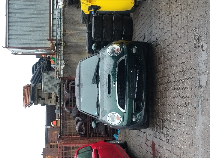 The scene depicts an outdoor industrial or commercial yard in Saarlouis, Germany, under an overcast sky with visible moisture on all surfaces, indicating rain or recent precipitation. The ground is covered with wet, grey interlocking paving stones. A dark green Mini Cooper (R56 generation), featuring a hood scoop, is centrally parked, facing the observer. Its exterior surfaces are visibly wet. To the left of the Mini, the front portion of a red compact car is partially visible, also appearing wet. In the background, to the left, large reddish-brown metal cages filled with numerous used tires are stacked. Behind these cages, a dense accumulation of industrial materials, including concrete blocks, corrugated metal sheets, and black pipes, is evident. A concrete retaining wall spans horizontally across the mid-ground. To the right of the Mini, a tall stack of black tires is positioned next to two bright yellow recycling bins, partially obscured. Further in the background, a large, silver-colored corrugated metal skip container or structure is present. No individuals are visible, and no actions or interactions are occurring.