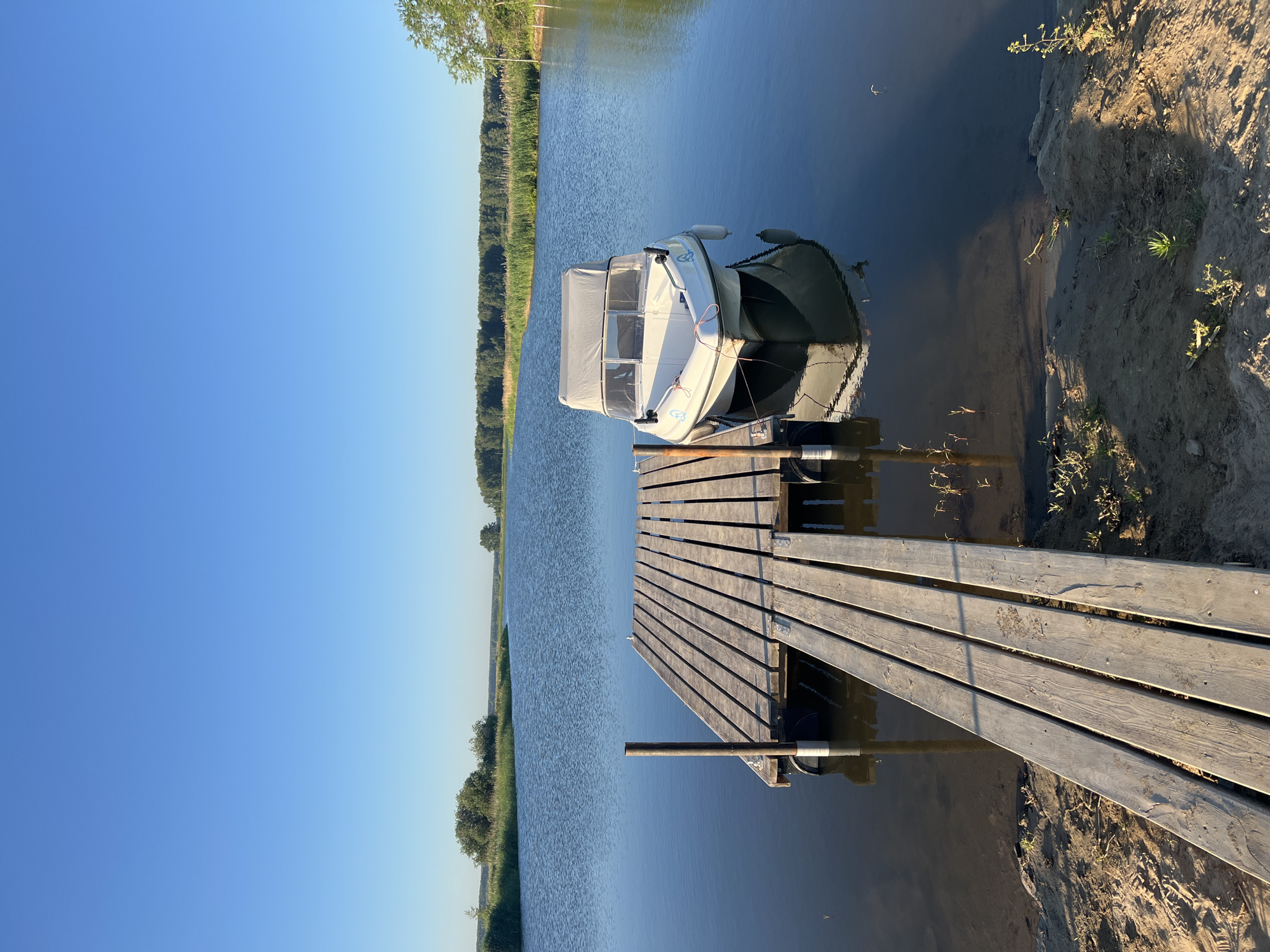 The image depicts a serene daytime scene at a waterfront in Novopichugovo, Russia. A wooden pier extends into a calm body of water, where a small white motorboat with a gray canopy is docked. The boat is tethered to the pier, and its reflection is visible on the water's surface. The pier is constructed of weathered wooden planks and has metal supports. The surrounding environment features lush green vegetation along the water's edge and a clear blue sky. The shoreline is sandy with sparse growth of grass and small plants. The overall atmosphere is peaceful and tranquil.