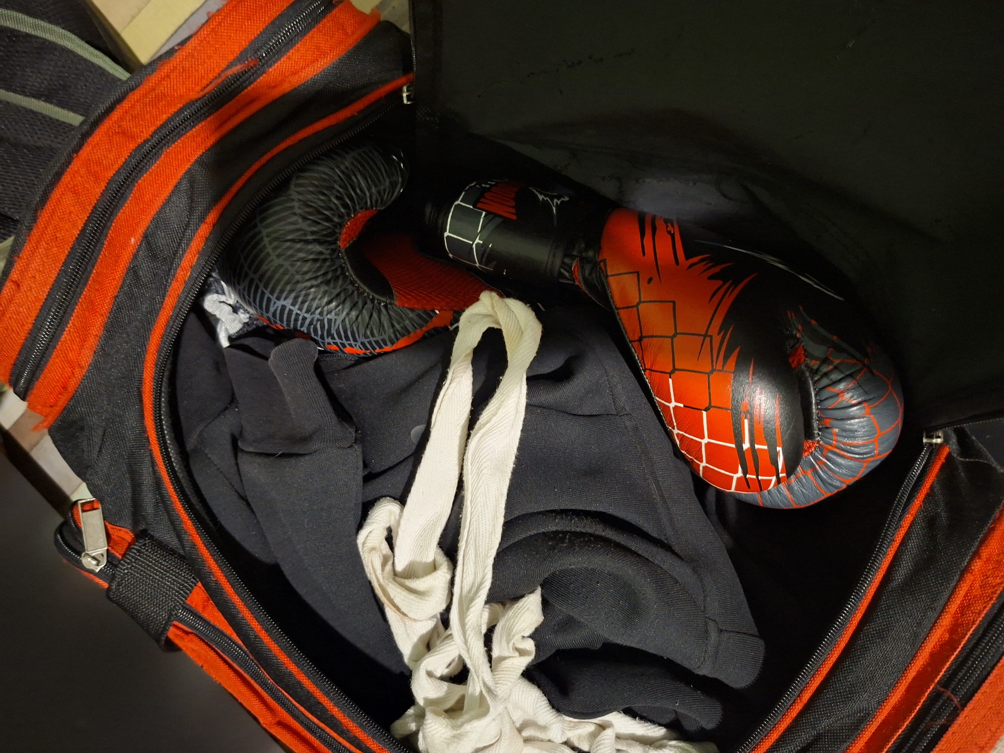 An overhead, close-up shot captures the interior of an open sports duffel bag. The bag itself is black with vibrant red stripes and zippers running along its edges. Inside, a pair of boxing gloves, predominantly black and red with a distinct grid pattern and stylized black scratch marks on the red sections, is visible. The left glove appears more black/grey with red accents, while the right glove is largely red with black accents and the patterned surface.
Beneath and around the gloves, there is a mass of dark, possibly black, fabric, which appears to be clothing, such as a shirt or shorts. A length of white, braided fabric, resembling hand wraps or shoelaces, lies draped over the black clothing near the center of the bag's opening.
The contents suggest preparation for or completion of a sports activity, likely boxing or martial arts training. The background outside the bag is dark and out of focus, providing no specific environmental cues about the location or time of day. No people or legible text are visible in the frame. The location context is Basarbovo, Bulgaria.