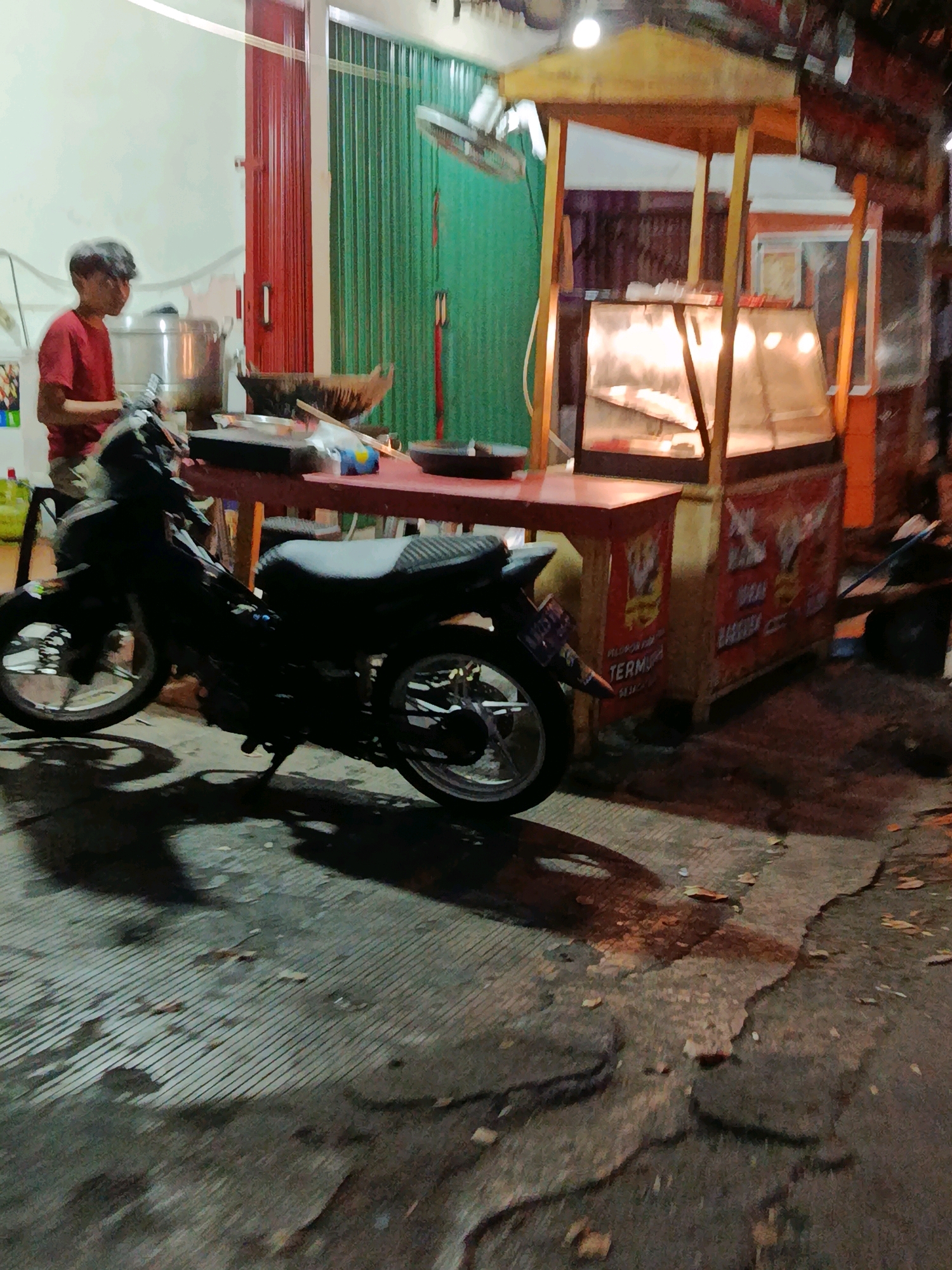 A young man in a red t-shirt stands near a food stall at night in a street in Bekasi, Indonesia. He is preparing food on a portable stove, with a large pot and other cooking utensils visible. A motorcycle is parked in front of the stall, casting a shadow on the wet, cracked pavement. The food stall is made of wood and has a glass display case for food, illuminated by lights. The area appears to be a street market or roadside eatery, with the overall scene suggesting a bustling commercial environment.