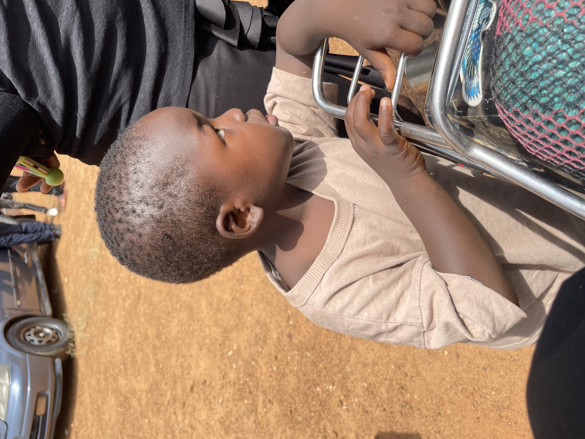 A young child with dark skin and short, dark hair is shown in a close-up profile view, looking to the right. They are wearing a light brown t-shirt and their hands are resting on a metal frame. The background shows a dusty, orange-brown surface, with a car visible on the left. The scene appears to be outdoors in an urban or semi-urban environment, possibly in Kassa, Nigeria, given the earthy tones and the presence of a vehicle. The lighting suggests it is daytime. The child's expression is neutral, and there is no clear interaction with other individuals or objects beyond their immediate grasp of the metal frame.
