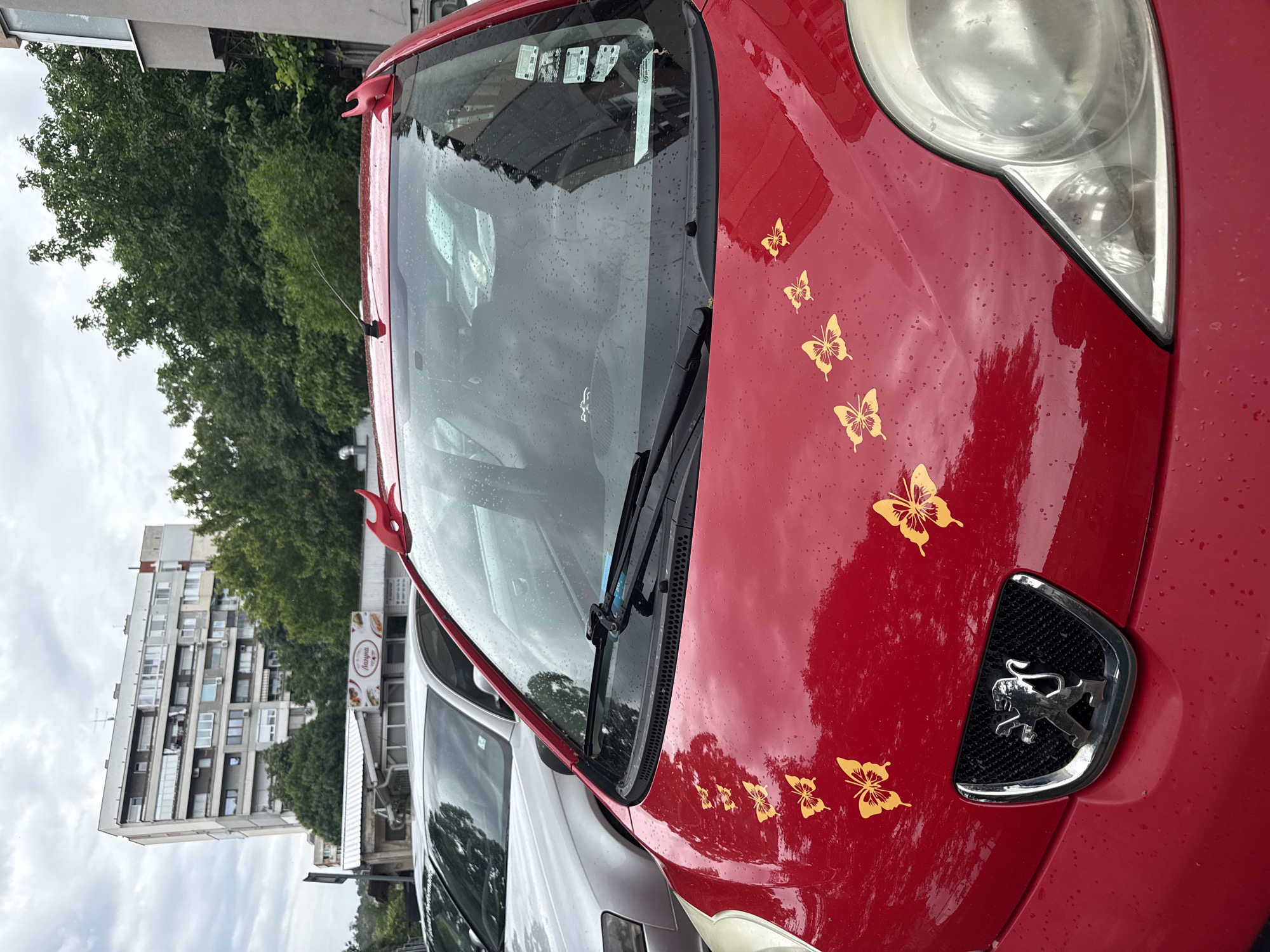 The image captures the front section of a red Peugeot car parked outdoors in what appears to be an urban setting in Plovdiv, Bulgaria, under an overcast sky. The car's hood features several yellow butterfly decals, and red, horn-like accessories are visible on its roofline near the windshield. Small water droplets cover the car's surface and windshield, indicating recent rain or mist. The distinctive Peugeot lion logo is prominently displayed on the car's grille.
In the background, a large, multi-story residential building with numerous windows and balconies rises, partially hidden by lush green trees. Another car, light-colored, is partially visible parked next to the red car. A street sign with the word "Багерия" (Bageriya), meaning "Bakery" in Bulgarian, along with images of baked goods, is visible in the mid-ground, suggesting the presence of a bakery nearby. The time of day appears to be daytime, given the ambient light, despite the cloudy conditions.