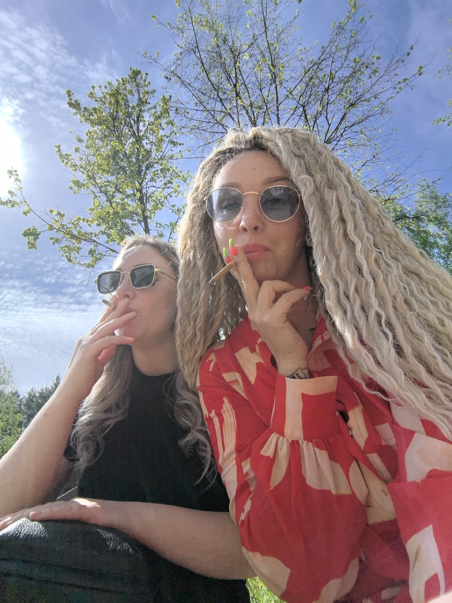 Two women are smoking outside on a sunny day. The woman on the right has long, blonde dreadlocks, sunglasses, and a red and white patterned shirt. She is holding a cigarette up to her lips and has brightly colored nail polish. The woman on the left has blonde, wavy hair, sunglasses, and a black shirt. She is also smoking a cigarette. In the background, there is a blue sky with white clouds and trees with green leaves. The location is Chaikovsky, Russia.