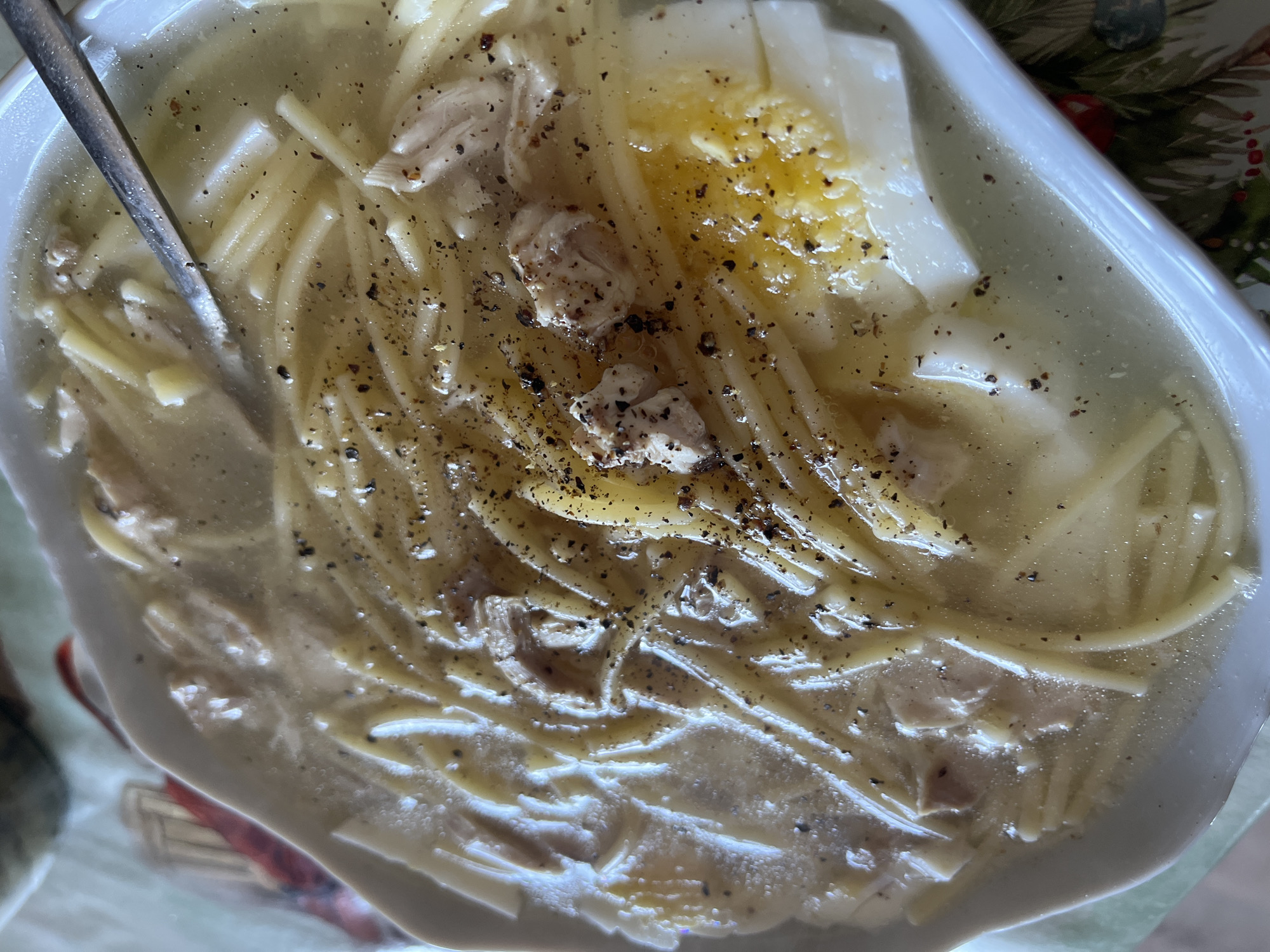 The image shows a bowl of chicken noodle soup with a boiled egg on top, sprinkled with black pepper. A spoon rests in the soup. The soup is served in a white bowl. The background shows a table with a Christmas-themed tablecloth. There is no evidence in the image to suggest it was taken in Chaikovsky, Russia.