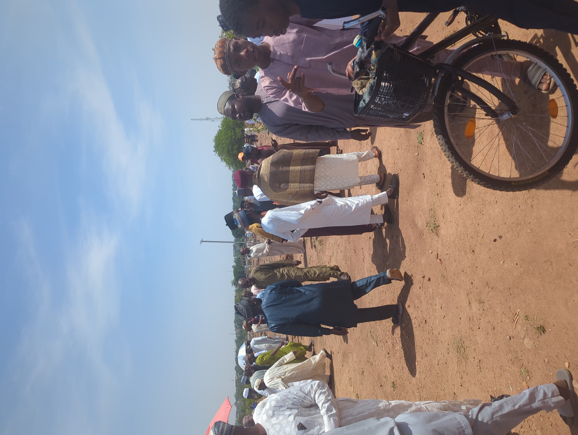 The image shows a group of men standing on a dirt ground under a clear sky. The men are mostly wearing traditional clothing, including robes and caps. A bicycle with a basket is visible on the right. The location is presumed to be in Sardauna, Nigeria, based on the user prompt. The men seem to be engaged in a conversation or gathering. A utility pole or tower is visible in the background.