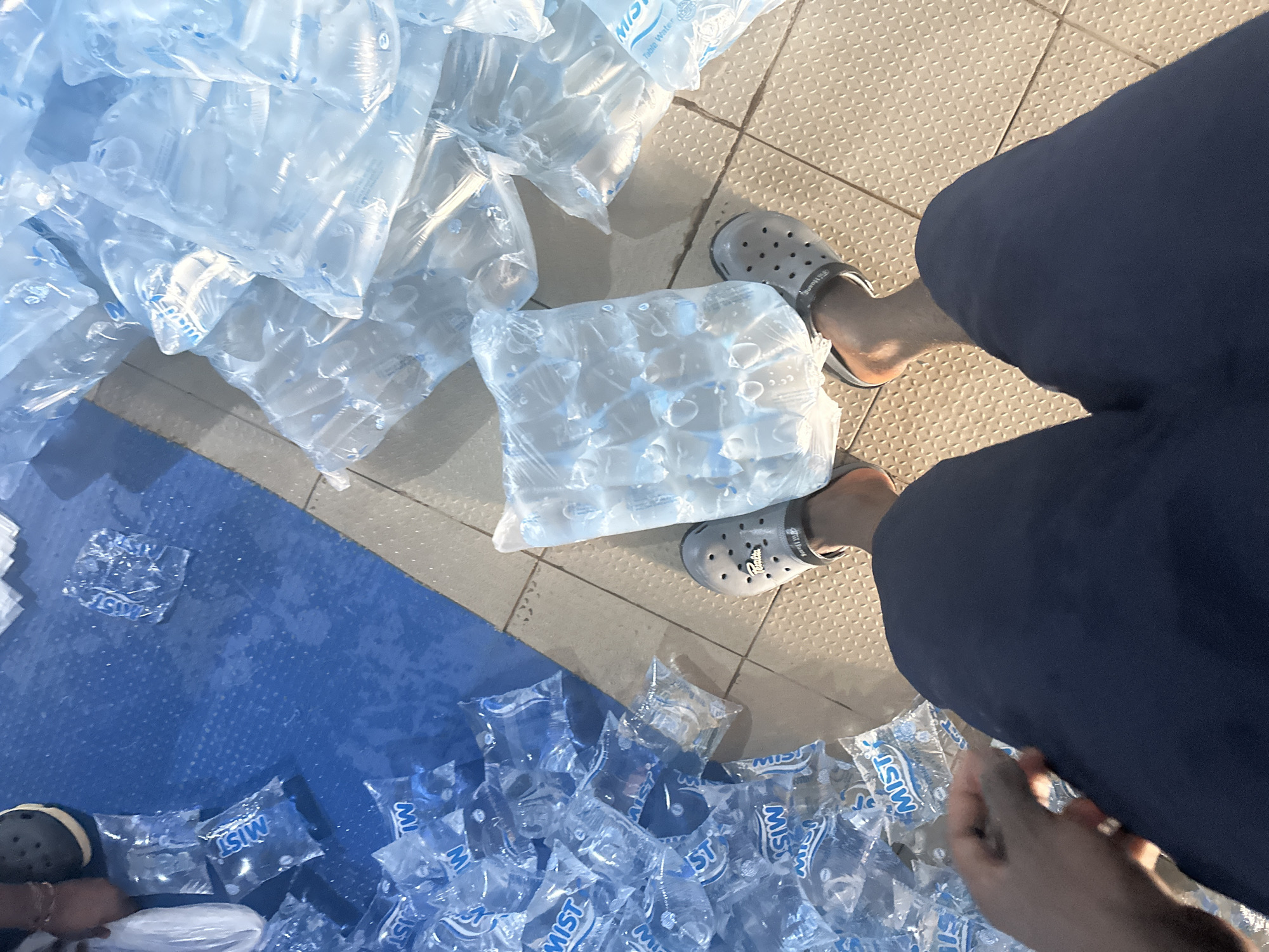The image presents an overhead view of a setting filled with numerous clear plastic sachets of drinking water, likely in an indoor or covered commercial space in Jos, Nigeria.
The main objects are countless individual water sachets, many of which are bundled together in larger transparent plastic bags, forming stacks and piles. Several loose sachets are scattered across the floor, suggesting handling or distribution. A recurring brand name "MIST" is clearly visible on the sachets, often accompanied by "Table Water."
Two individuals are partially visible from a high angle, primarily their lower bodies and feet. In the foreground, one person wears dark blue trousers and light grey Crocs-style sandals; their right hand is also seen. On the bottom left, another person's left foot, also in a similar grey sandal, is partially visible. The presence of these individuals indicates involvement in an activity related to the water sachets.
The floor is characterized by two distinct areas: a light brown or beige tiled section with a raised, textured pattern, and a smooth, dark blue section, possibly a mat or painted floor, separated by a straight line. The lighting is soft and even, without strong shadows, implying an indoor setting or an overcast outdoor condition.
The overall scene suggests an event of commercial distribution, storage, or possibly preparation of aid packages involving "pure water" sachets, a common form of drinking water in Nigeria. The sandals worn by the individuals have legible text on their straps, possibly "Paladin." No strong emotions are discernible from the partial view of the people.