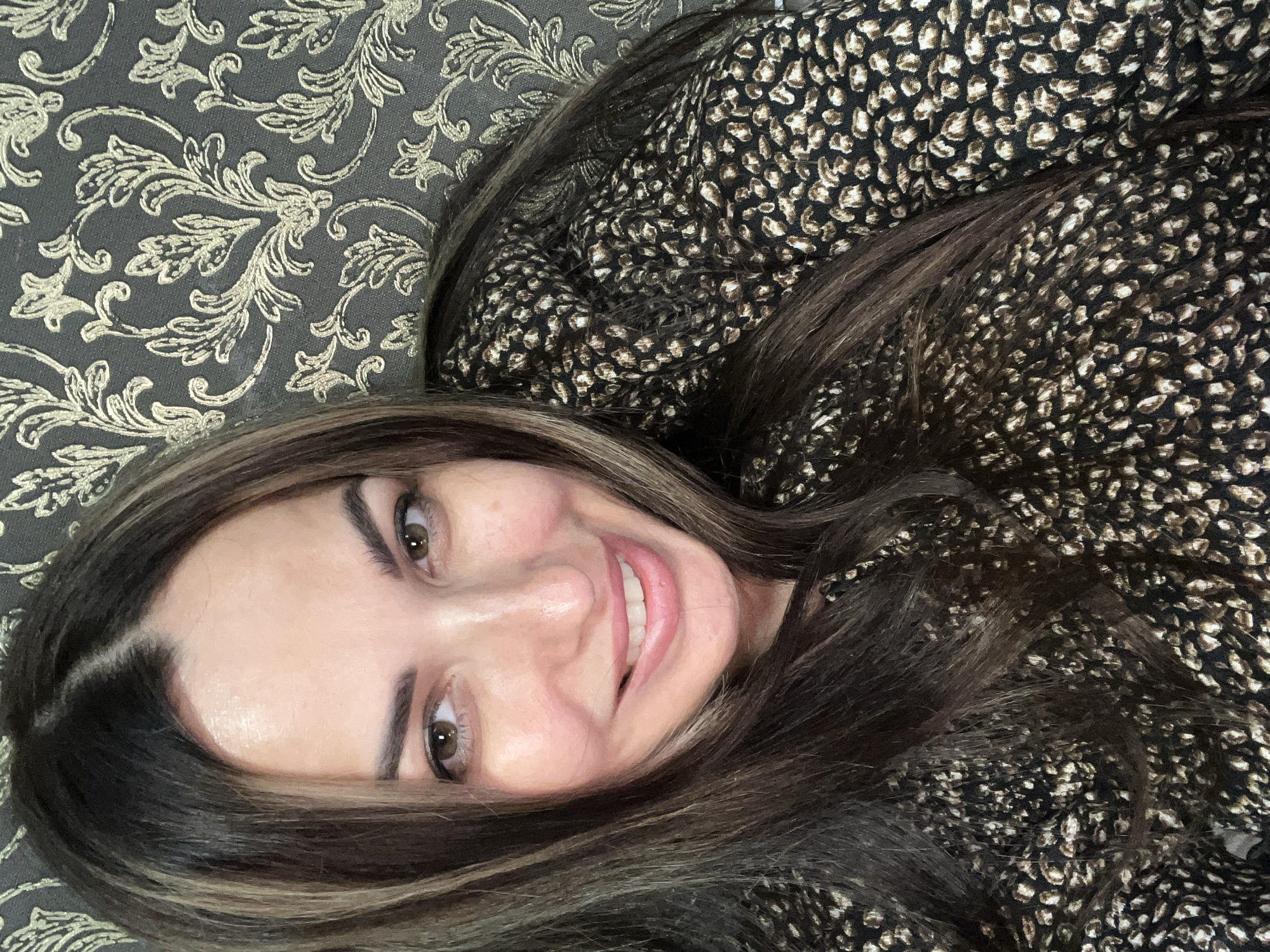 A woman with long, dark hair and highlights smiles at the camera, with her head tilted slightly to her left. She is wearing a black dress with a brown and white leopard-print pattern. The background consists of dark gray wallpaper with a repeating gold floral pattern. The lighting is soft and appears to be from a single source, illuminating her face and the foreground.