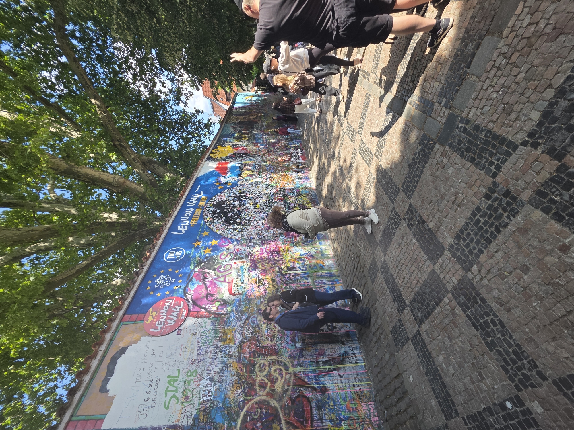 The image shows the Lennon Wall, a graffiti-covered wall in Prague, Czech Republic. A group of people are interacting with the wall, some photographing it, others standing and looking at the art. A couple is seen kissing in front of the wall. The wall is densely covered with graffiti, messages, and images, with the words "Lennon Wall" clearly visible. Trees cast shadows on the wall and surrounding area. The ground is paved with a pattern of dark and light stones.
