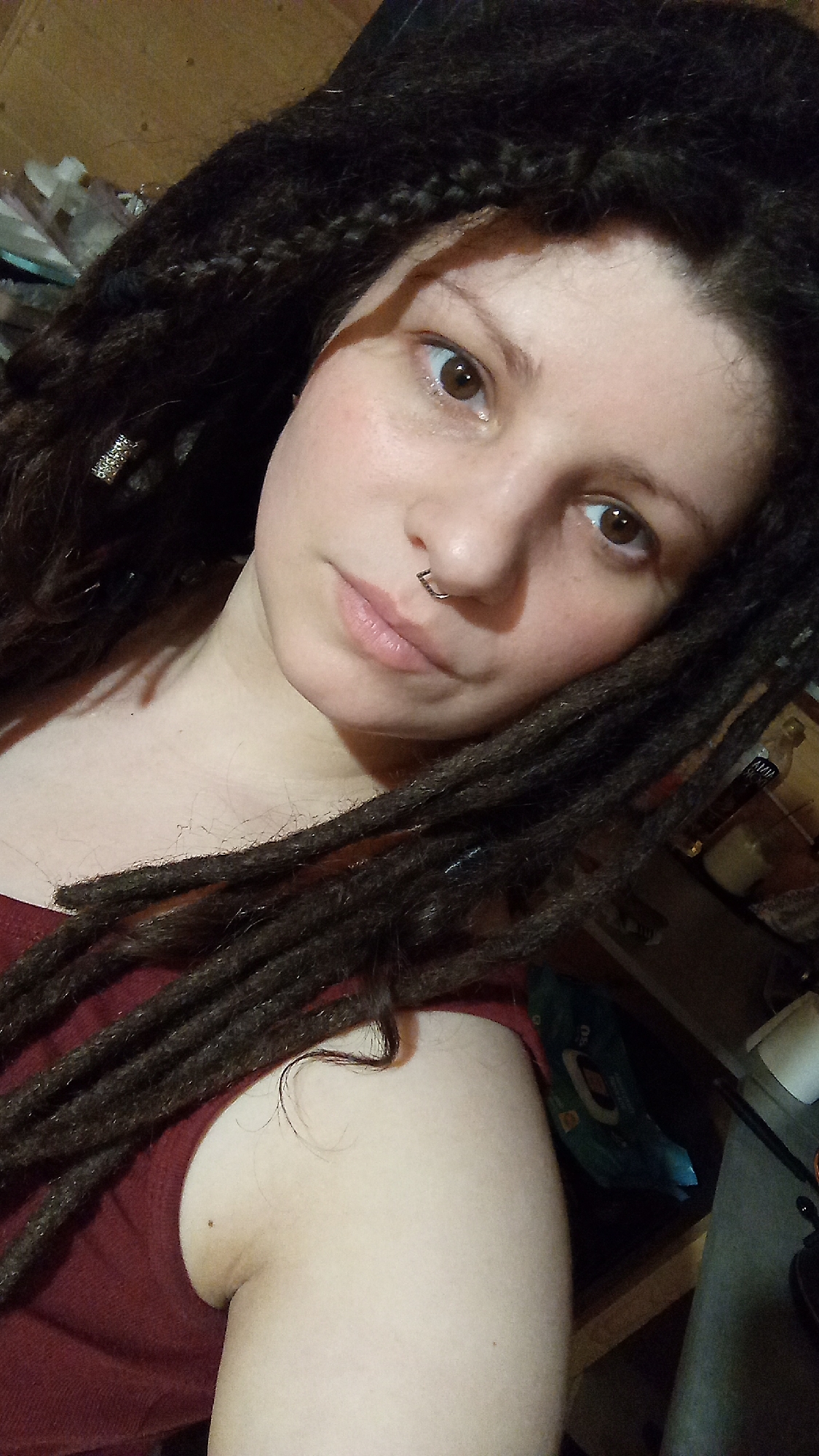 A young woman with brown eyes and a nose ring is shown in a close-up photograph. She has dark, dreadlocked hair with a decorative silver piece in one section. She is wearing a maroon top. The background is cluttered, with various items visible, including a bottle and wipes. The photo appears to have been taken indoors with ambient lighting. There is no evidence to suggest that the photograph was taken in Юдино, Russia.