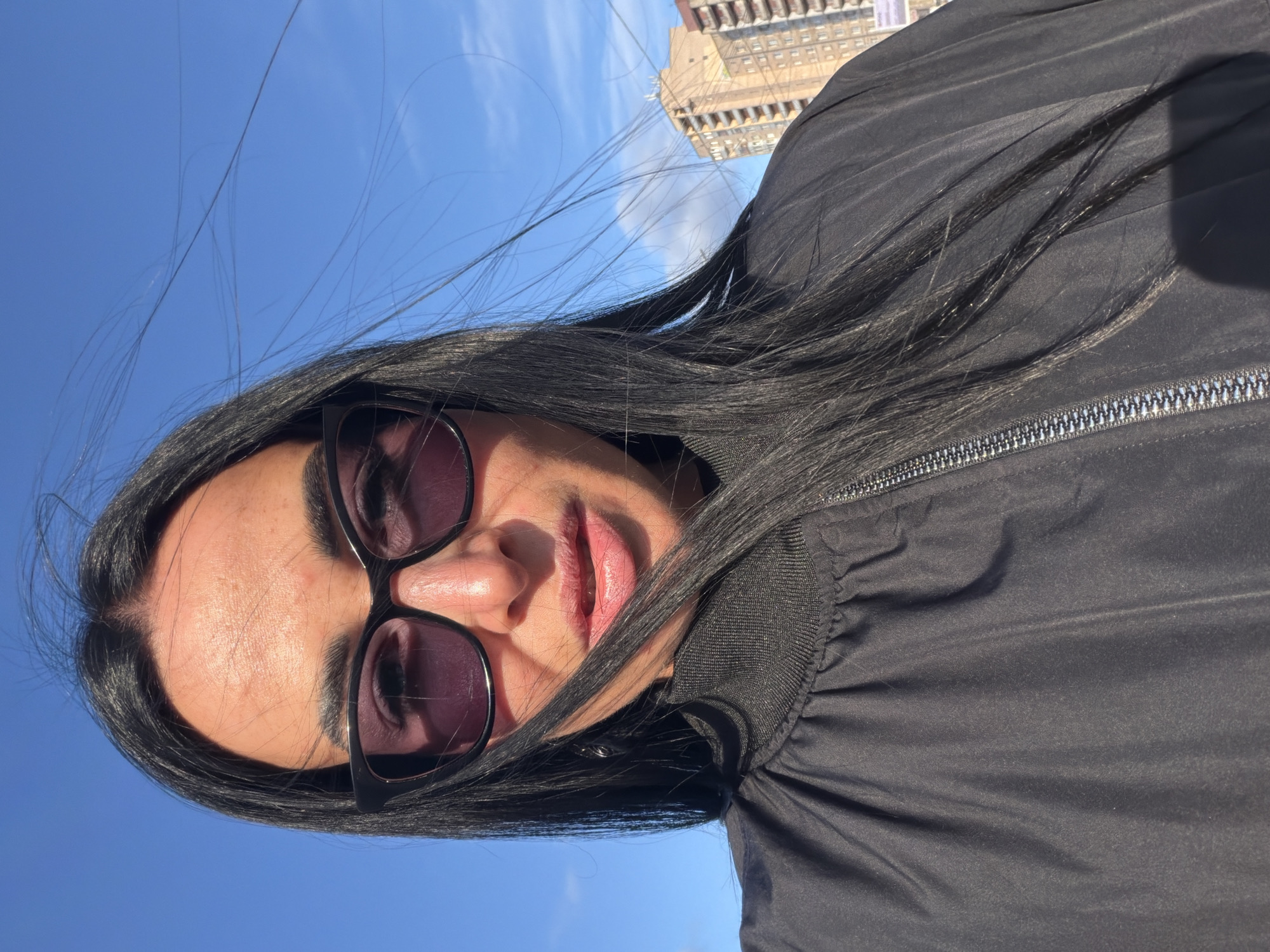 The image is a selfie taken by a person with long, black hair who is wearing sunglasses and a black jacket. In the background, the clear, blue sky is visible, as well as the Saint Petersburg Stock Exchange building. The person is looking directly at the camera. The photo was taken outdoors on a sunny day.
