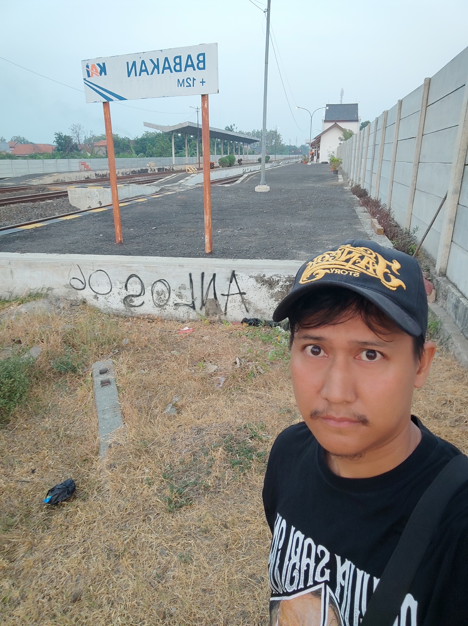 The image shows a man standing in front of a train station sign that reads "BABAKAN" and "MSR+". The station appears to be in a rural or semi-rural area, with dry grass and a concrete wall in the foreground. The man is wearing a black t-shirt and a black baseball cap with a gold logo. He is looking at the camera with a neutral expression. In the background, there are train tracks, a small building, and trees. The sky is overcast. The graffiti on the wall appears to be "20 20 14". The location is identified as the Babakan train station in Pakusamben, Indonesia.